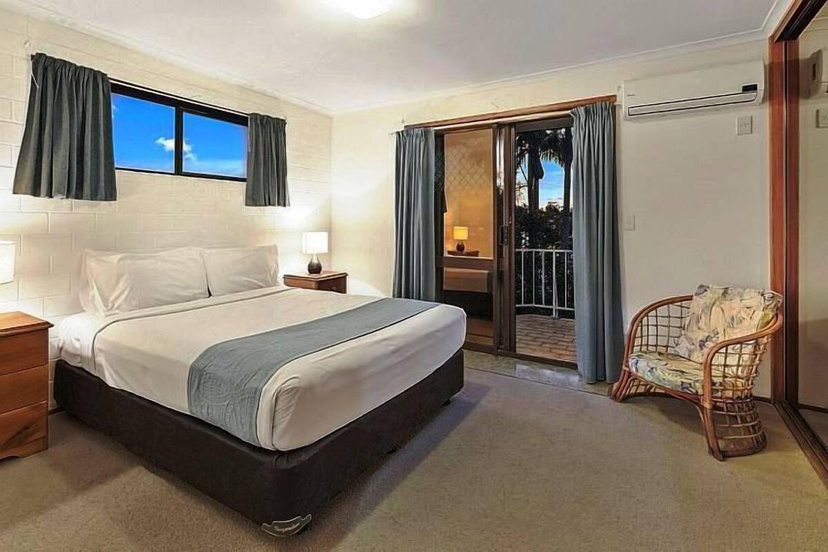 A bedroom with a bed, bedside tables, a chair, a window with curtains, and a sliding glass door leading to a balcony.