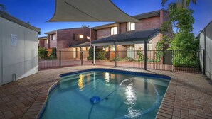 A rectangular swimming pool with a blue border, surrounded by a brick patio and a metal fence.