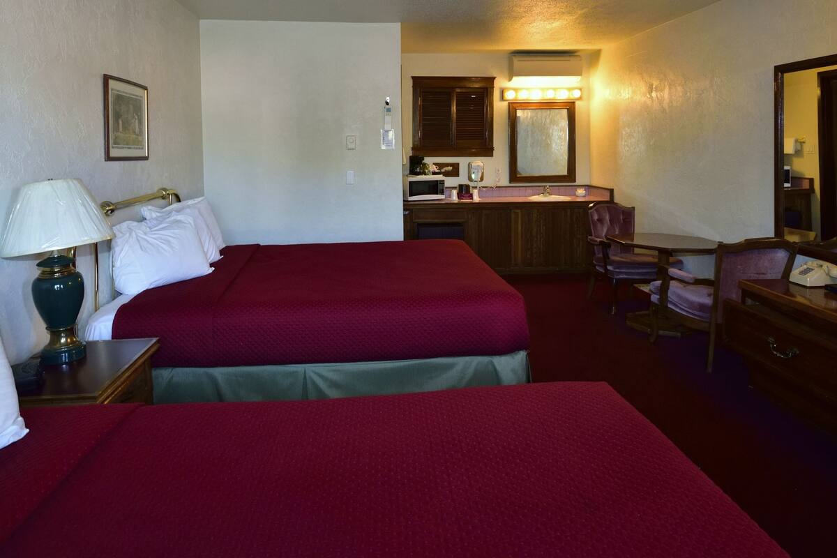 A hotel room with two beds, a desk with a lamp, a chair, a dresser, a television, and a bathroom with a sink and mirror.