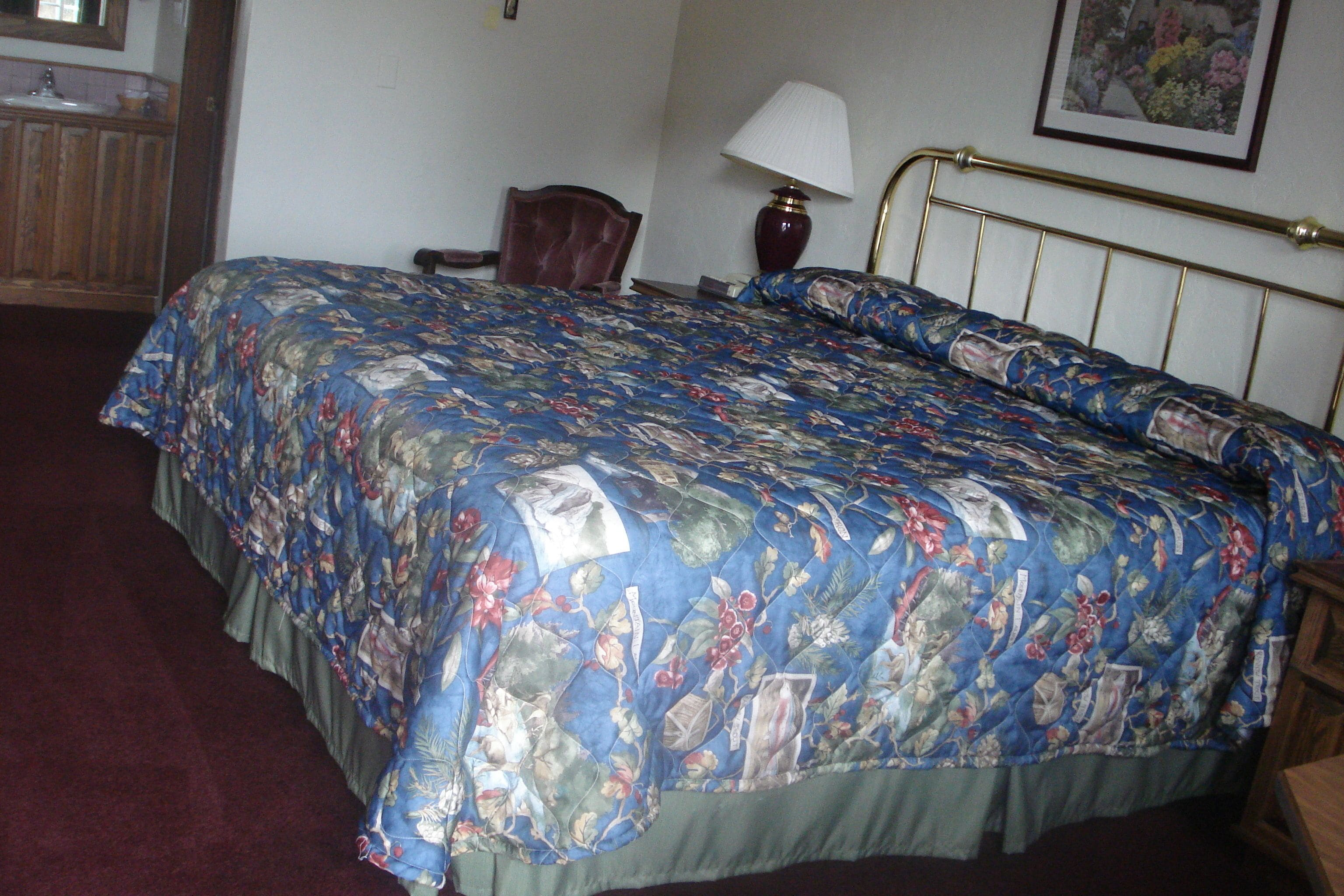 A bedroom with a floral bedspread, a wooden headboard, a bedside table, a lamp, and a framed picture on the wall.