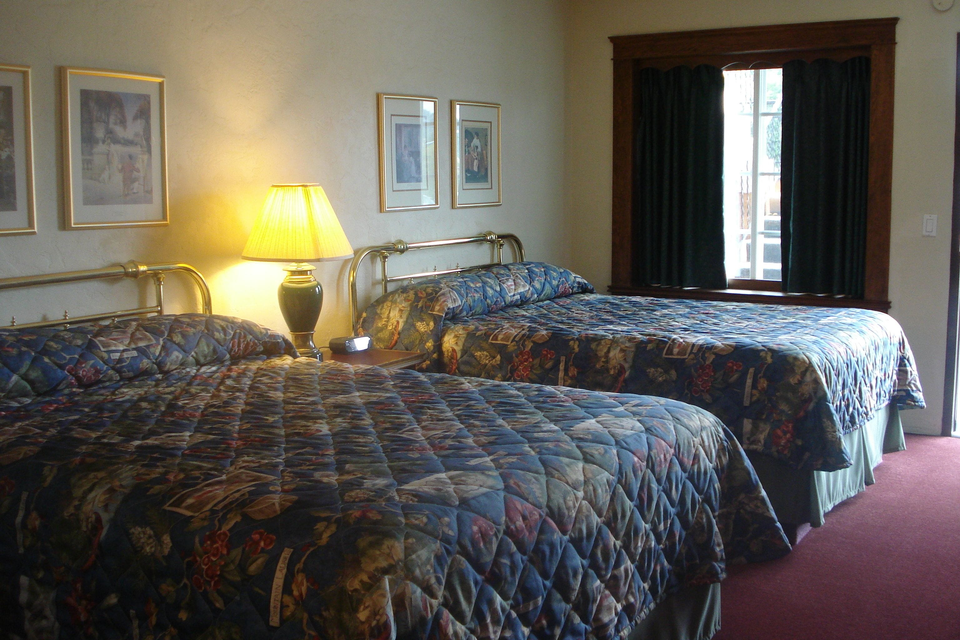 Ein Hotelzimmer mit zwei Betten, einer Lampe, gerahmten Bildern an der Wand und einem Fenster mit Vorhängen.