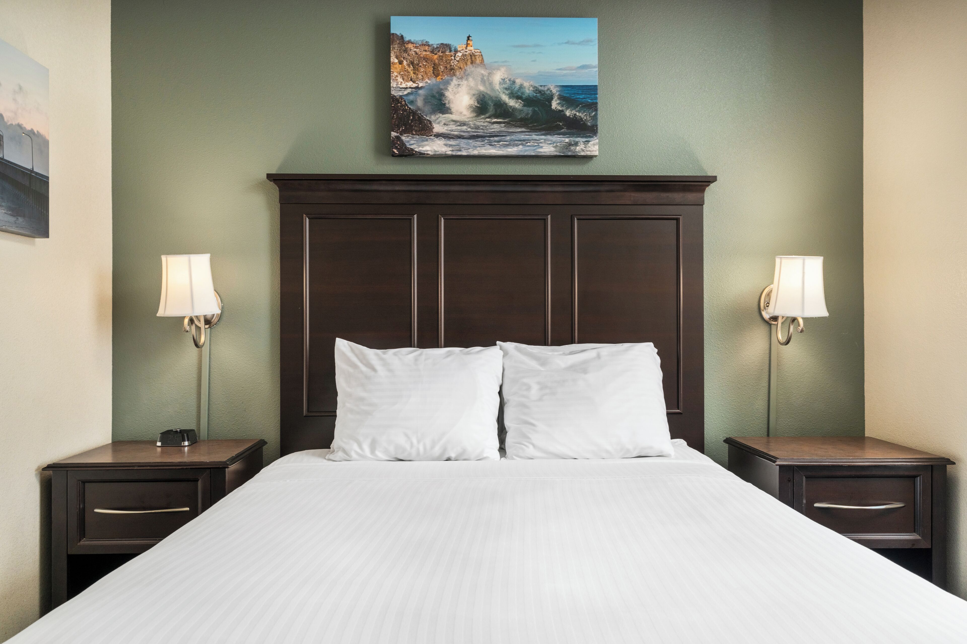 A hotel room with a large bed, two nightstands, and a painting of a coastal scene above the bed.