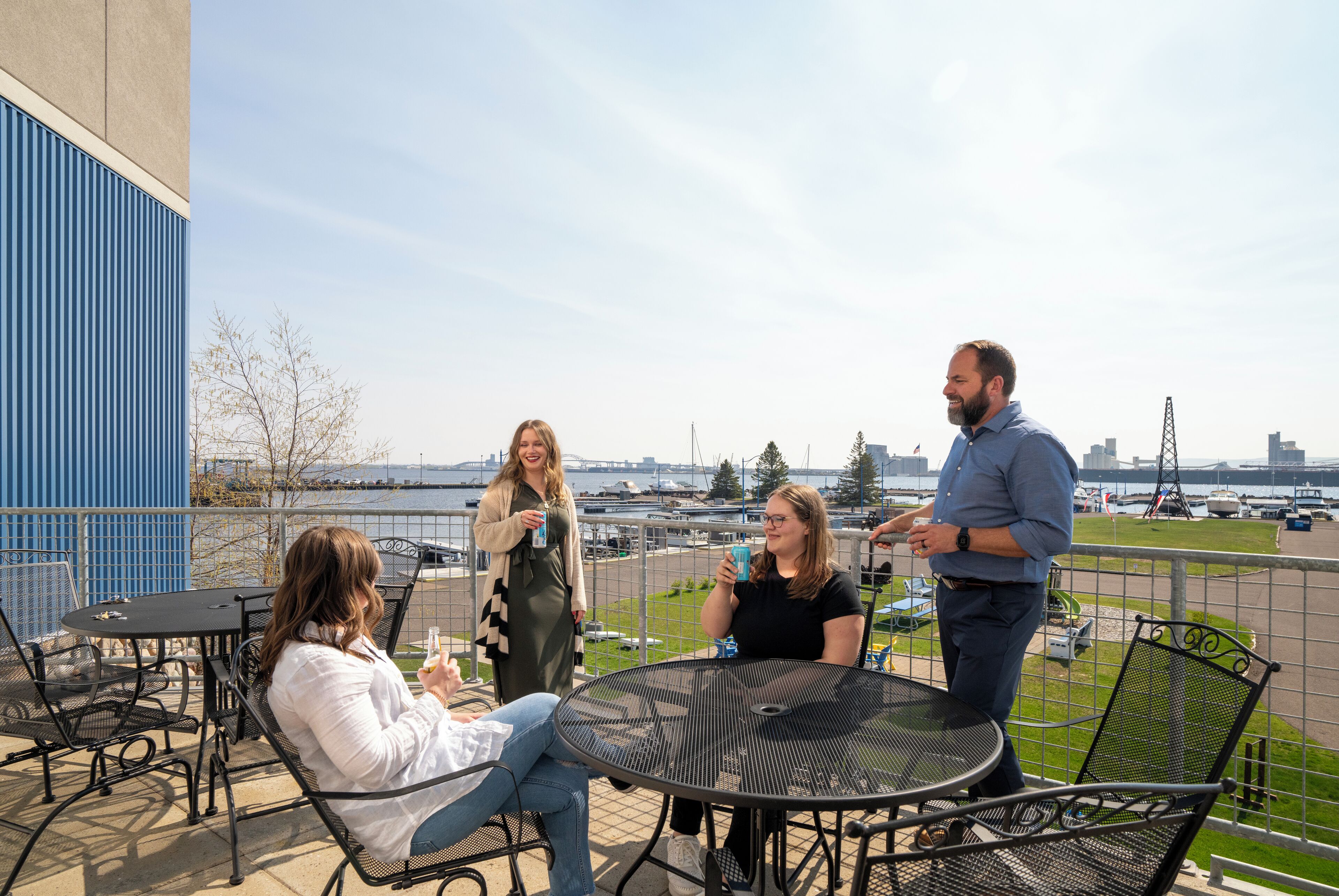 Un groupe de personnes est assis et se tient debout sur une terrasse en plein air, d’où l’on peut admirer la vue sur l’eau et le panorama de la ville.