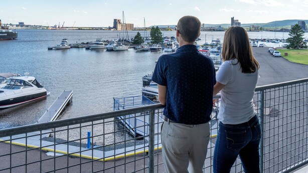 A couple overlooking a marina with boats docked, a bridge in the background, and a view of the cityscape.
