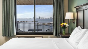A hotel room with a large window offering a view of a bridge and water. There is a bed, a nightstand with a lamp, and a vase with flowers. - Park Point Marina Inn (Duluth)