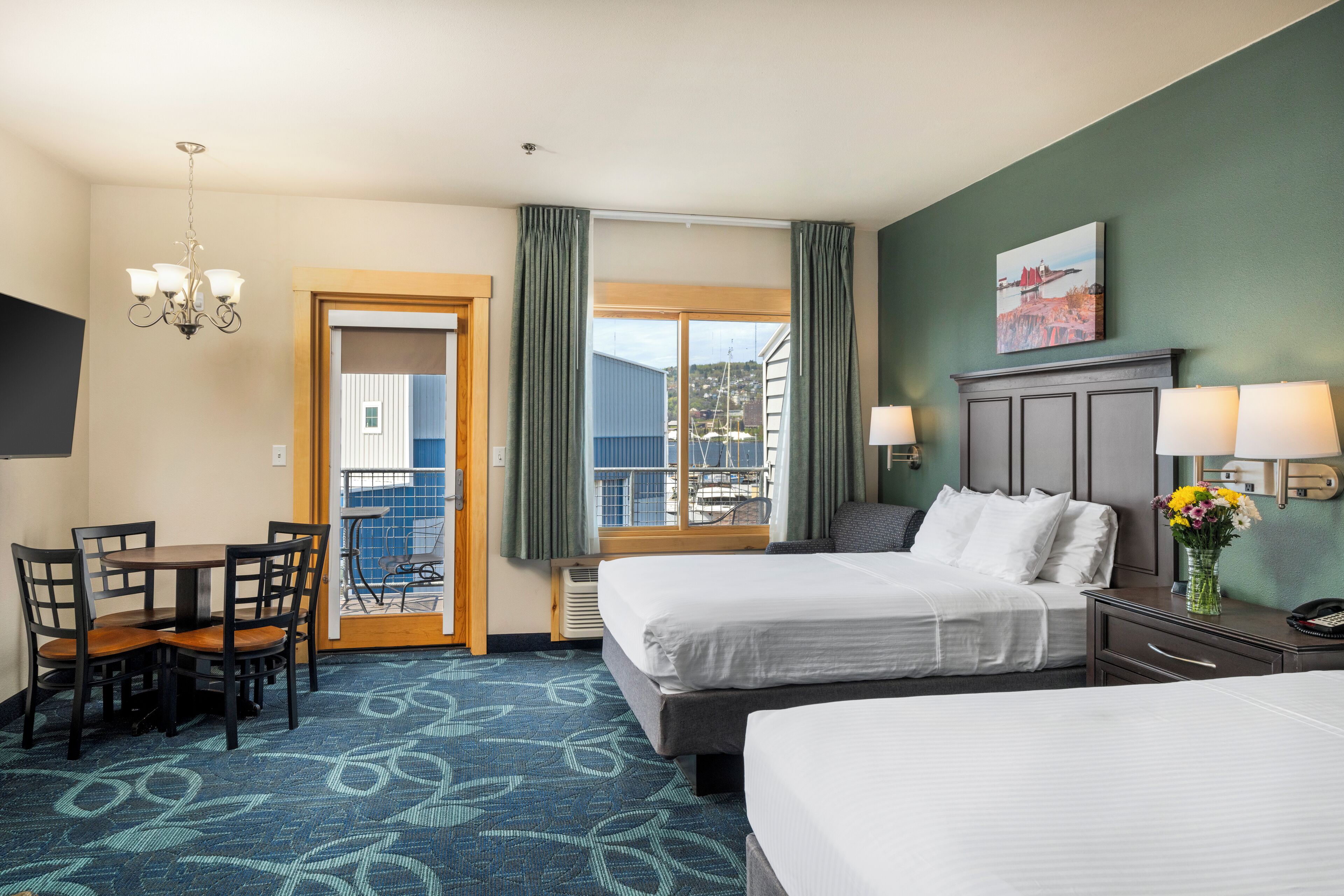 A hotel room with two beds, a desk with chairs, a chandelier, a window with curtains, a painting on the wall, and a view of boats and buildings outside.