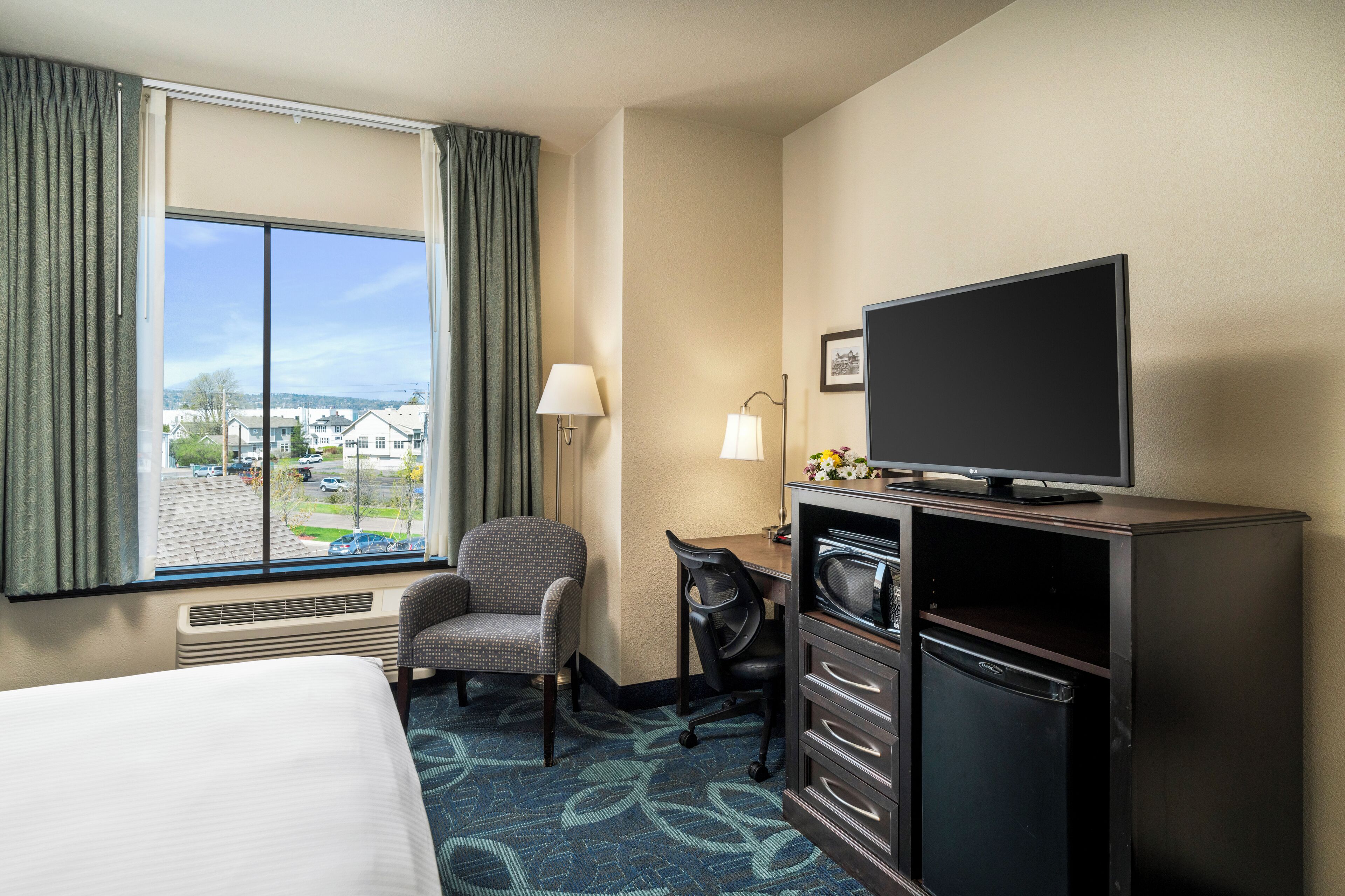 A hotel room with a bed, a desk with a chair, a television, a mini-fridge, and a window with curtains.