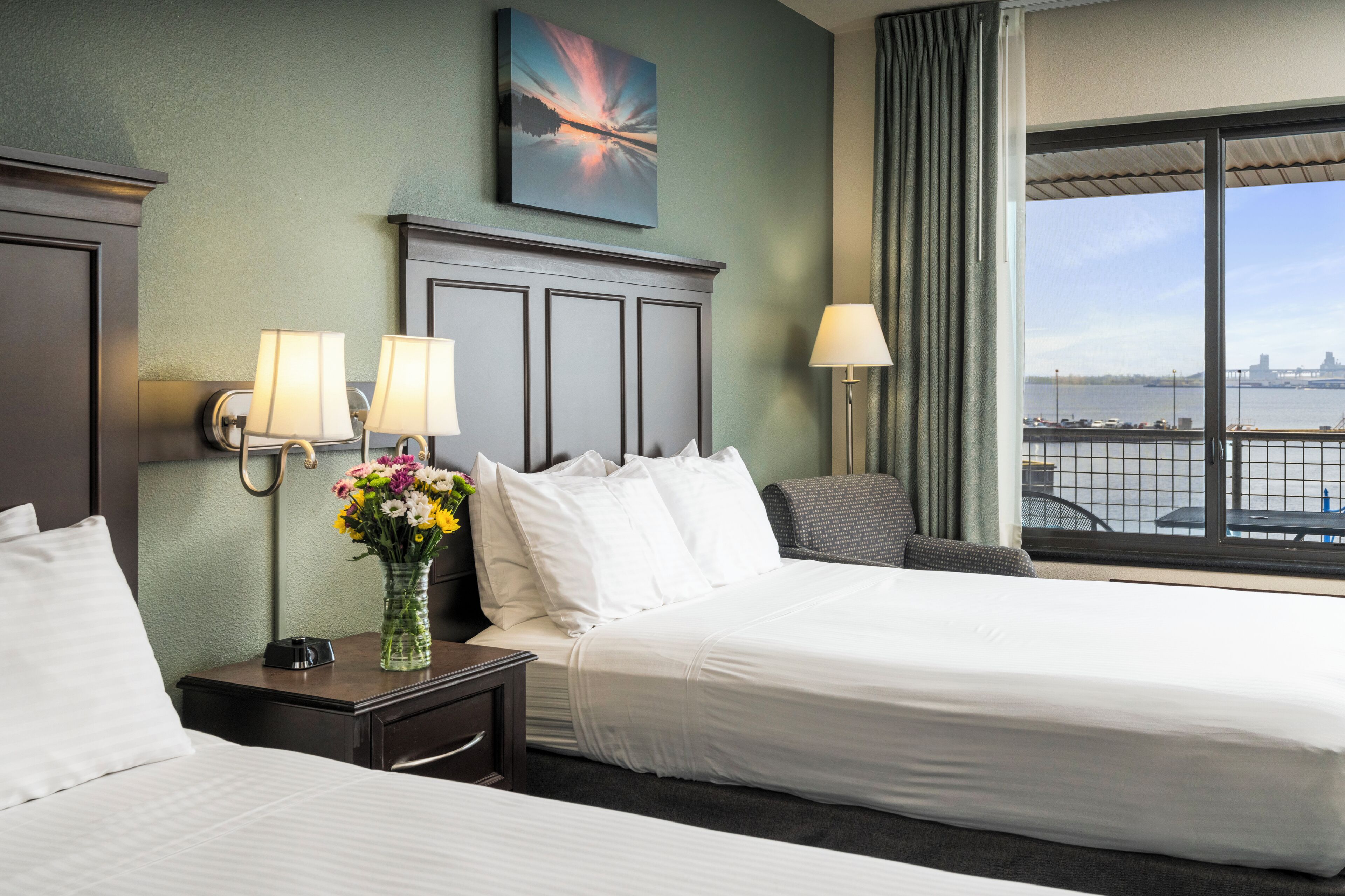 Habitación de hotel con dos camas, una mesita de noche con un jarrón de flores, un cuadro en la pared y vistas a un cuerpo de agua desde la ventana.