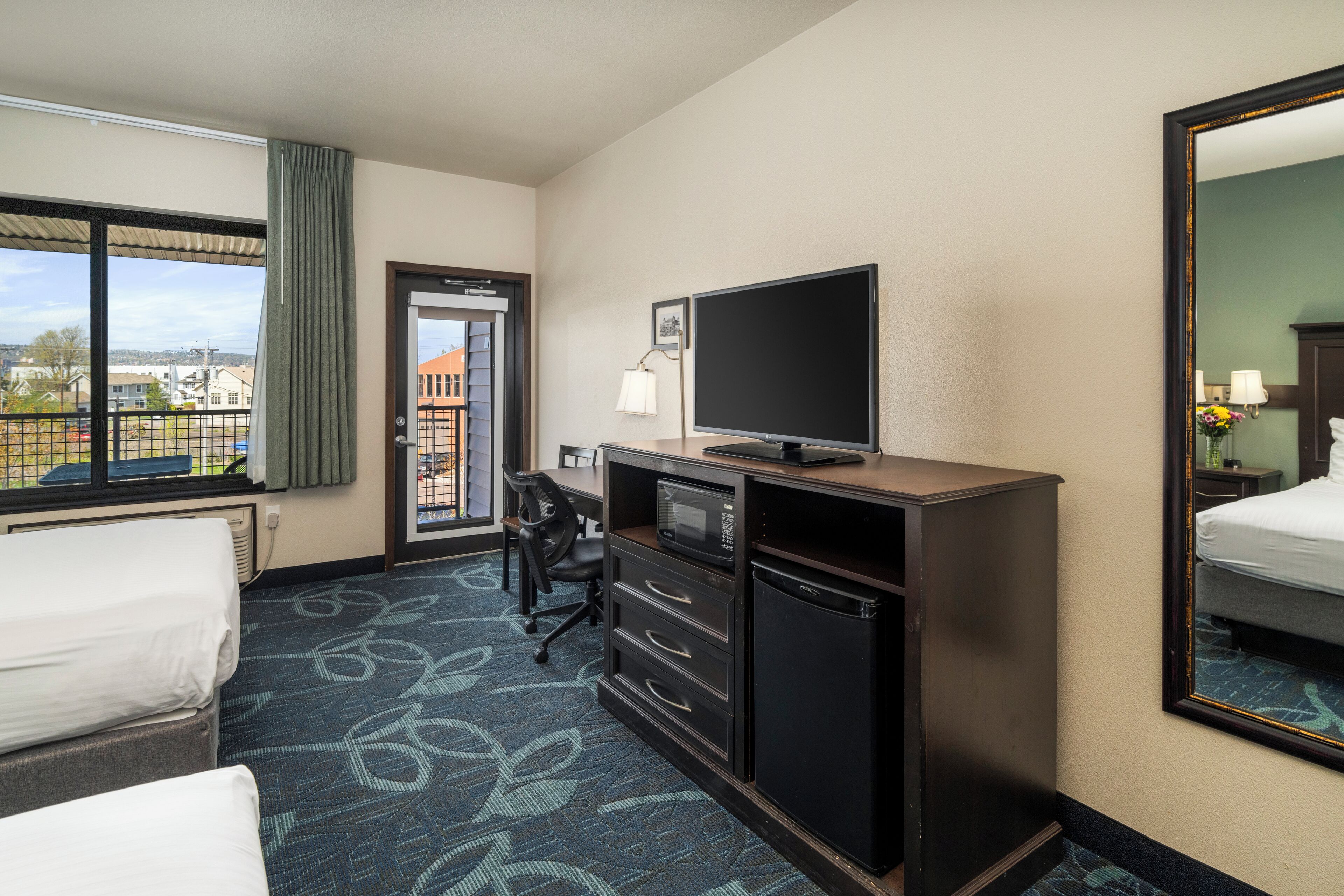A hotel room with a bed, a desk with a computer, a television, and a large window offering a city view.
