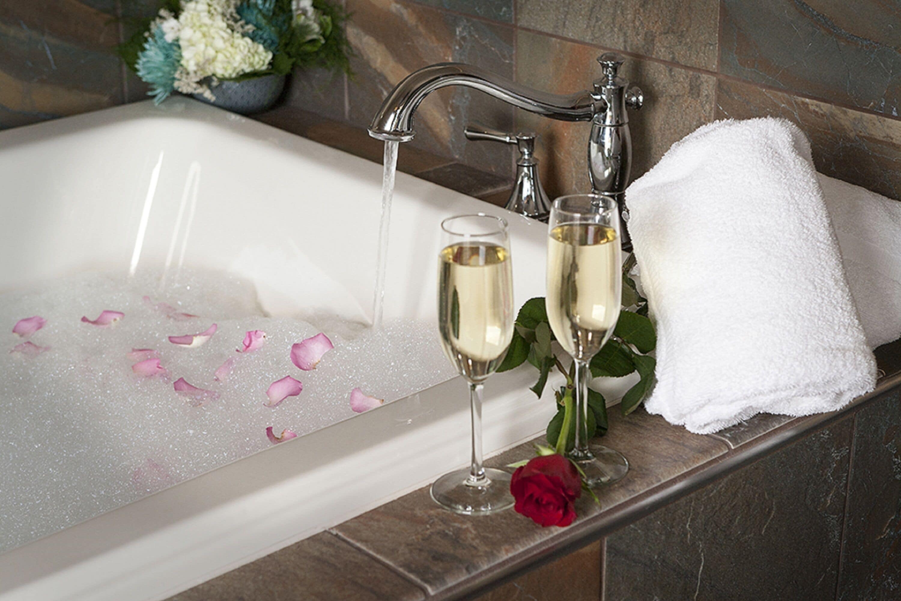 A bathtub with rose petals, two champagne glasses, and a white towel.