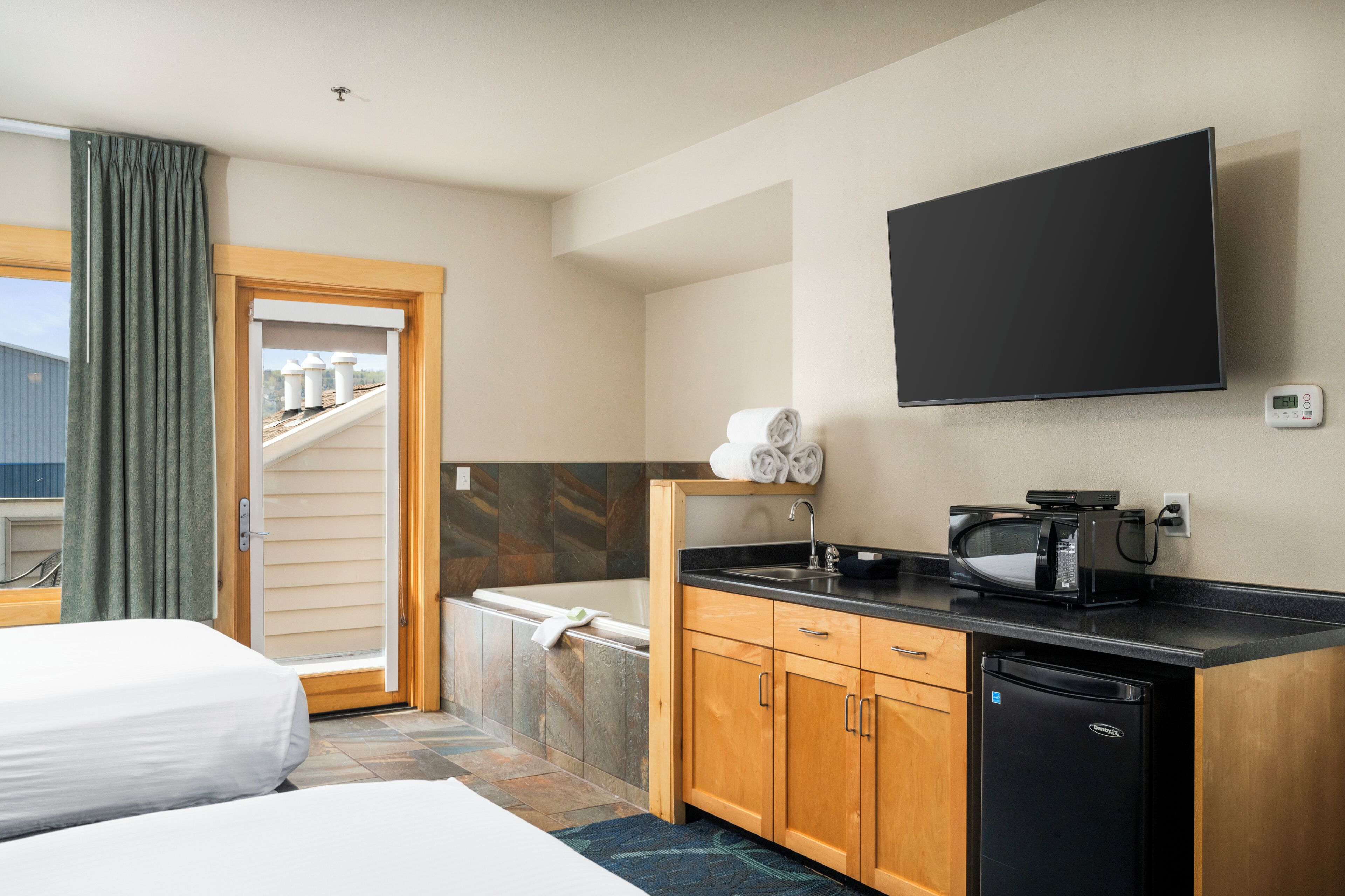 A hotel room with a bed, a bathroom with a bathtub, a kitchen area with a microwave, and a flat-screen TV mounted on the wall.