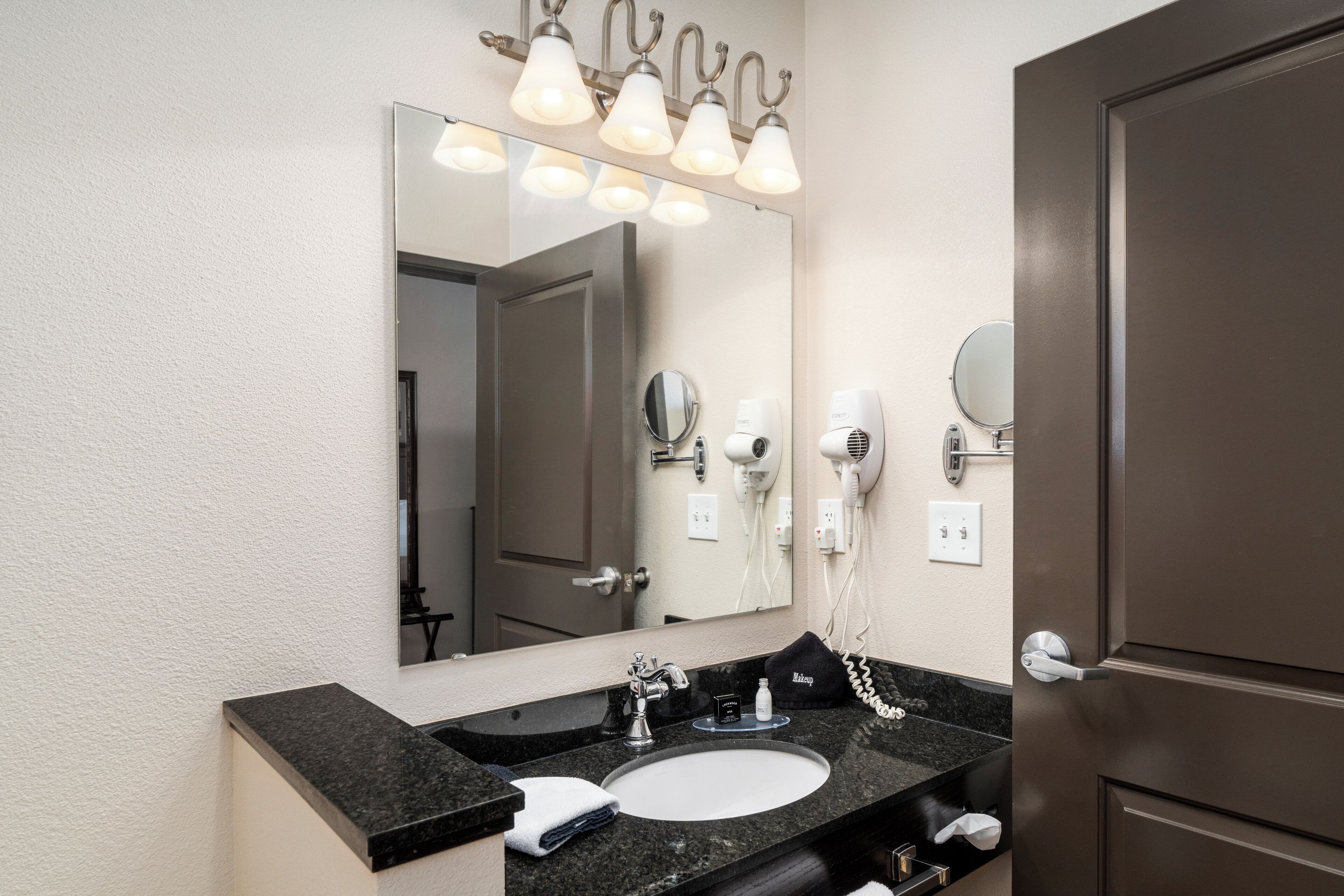 A hotel bathroom with a large mirror, a sink, a towel, and hair dryers on the wall.
