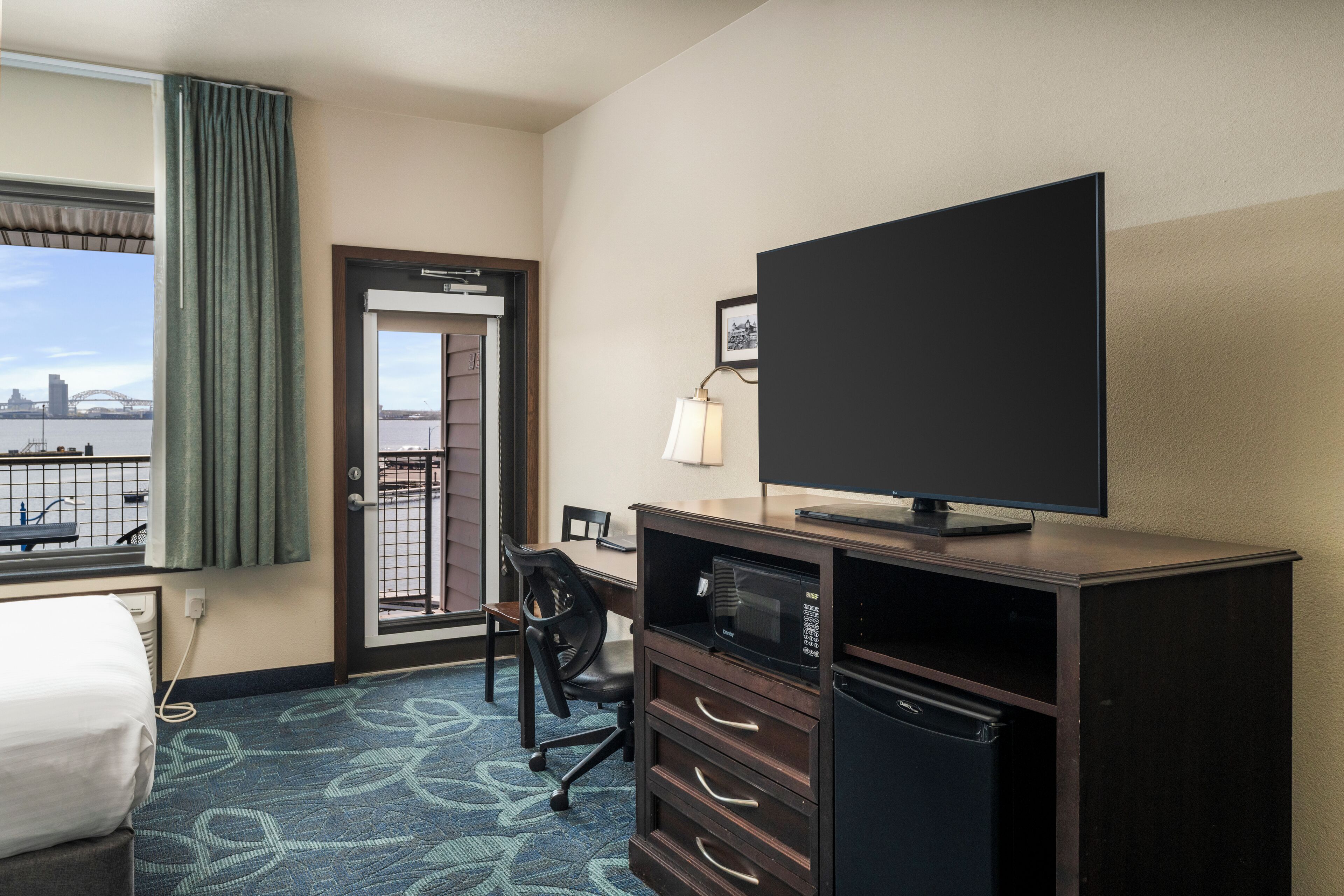 A hotel room with a bed, a desk with a computer, a television, a microwave, and a refrigerator. There is a window with a view of a bridge and a cityscape.