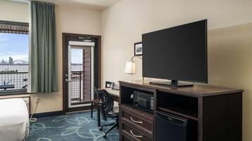 A hotel room with a bed, a desk with a computer, a television, a microwave, and a refrigerator. There is a window with a view of a bridge and a cityscape.