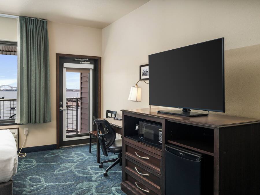 A hotel room with a bed, a desk with a computer, a television, a microwave, and a refrigerator. There is a window with a view of a bridge and a cityscape.