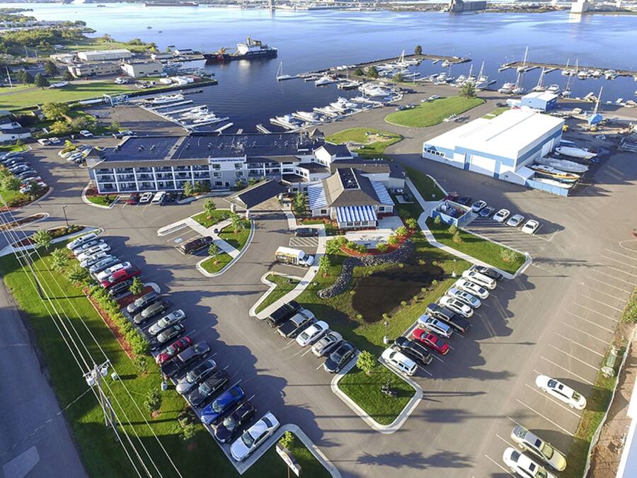 An aerial view of a marina with boats docked, a large building with a blue roof, and a parking lot with various vehicles.