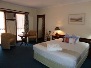 A hotel room with a bed, a chair, a small table, a lamp, and a window with curtains. - The Miners Rest Motel (Somerville)