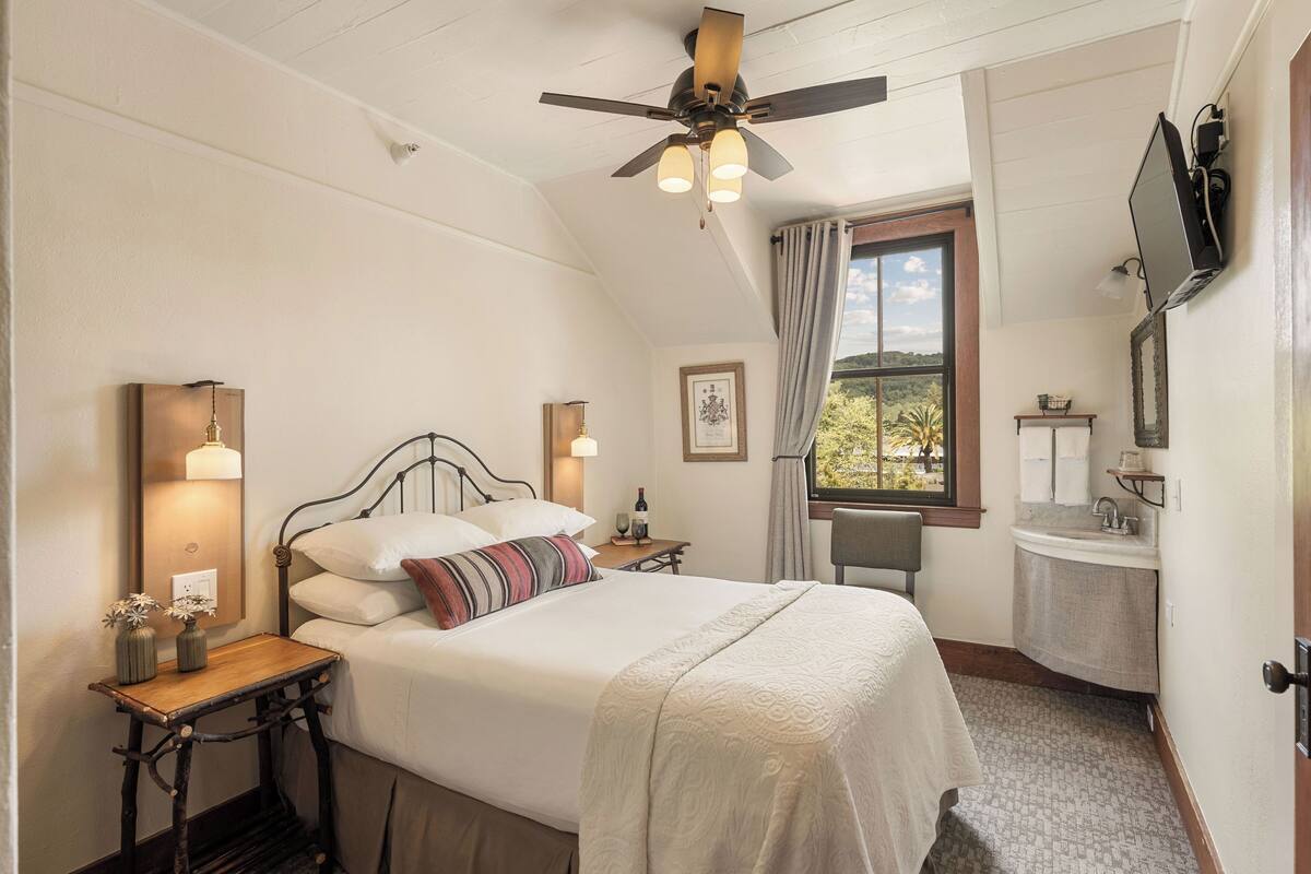 Une chambre à coucher comprenant un lit, des tables de chevet, un ventilateur de plafond, une fenêtre avec des rideaux, un téléviseur fixé au mur, et une salle de bain avec un lavabo et un miroir.