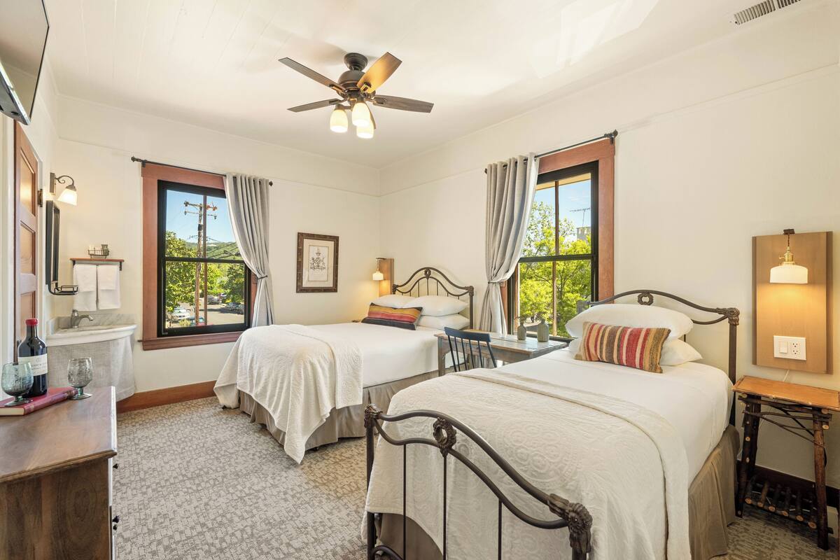 Une chambre avec deux lits, un ventilateur de plafond, une petite table avec une lampe et une fenêtre avec des rideaux.