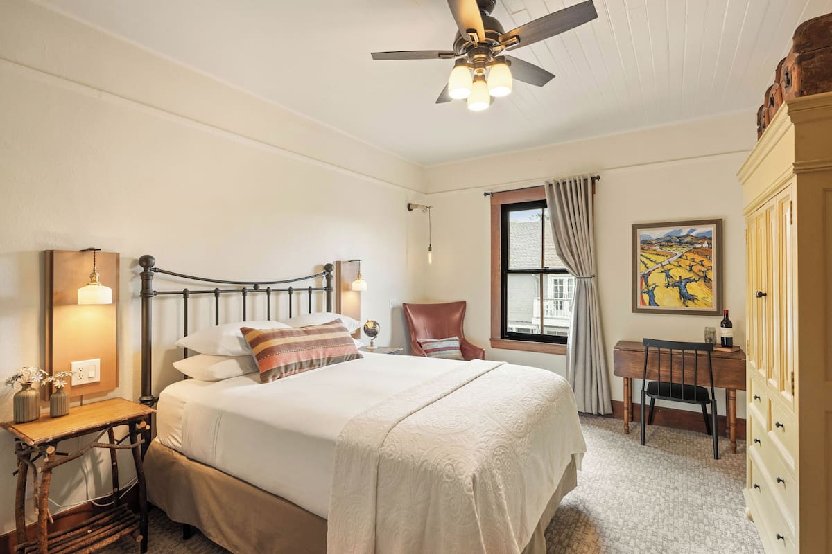 Une chambre à coucher comprenant un lit, un bureau, une chaise, un ventilateur de plafond et une fenêtre avec des rideaux.