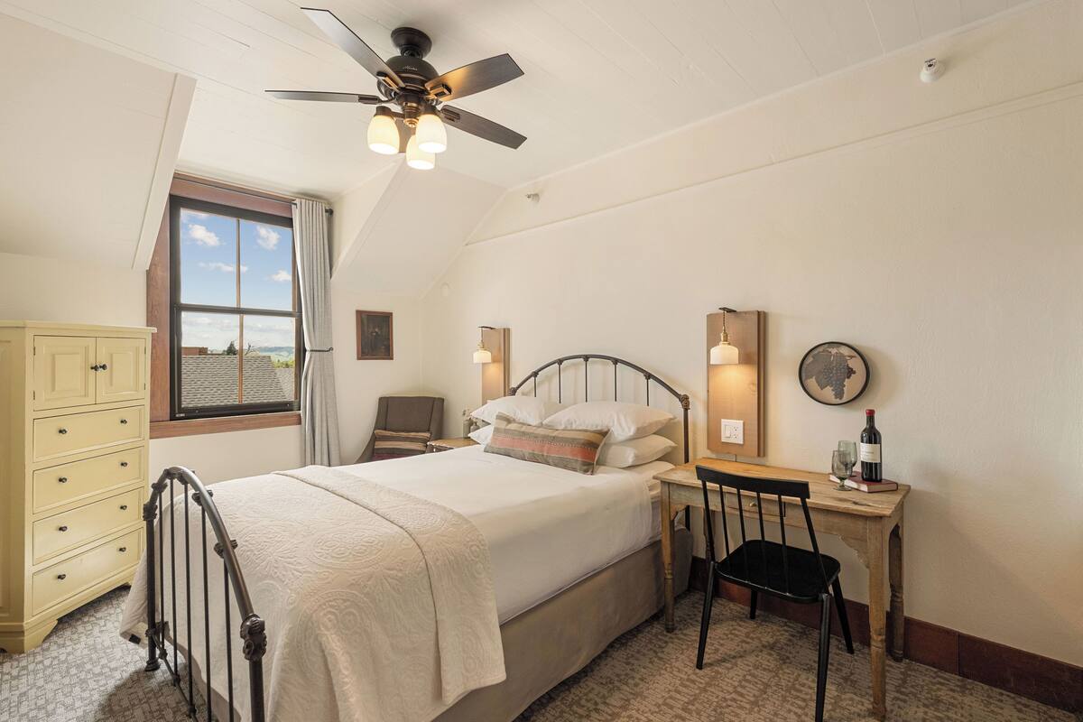 Une chambre à coucher comprenant un lit, un bureau, une chaise, une armoire, un ventilateur de plafond et une fenêtre donnant sur un beau paysage.