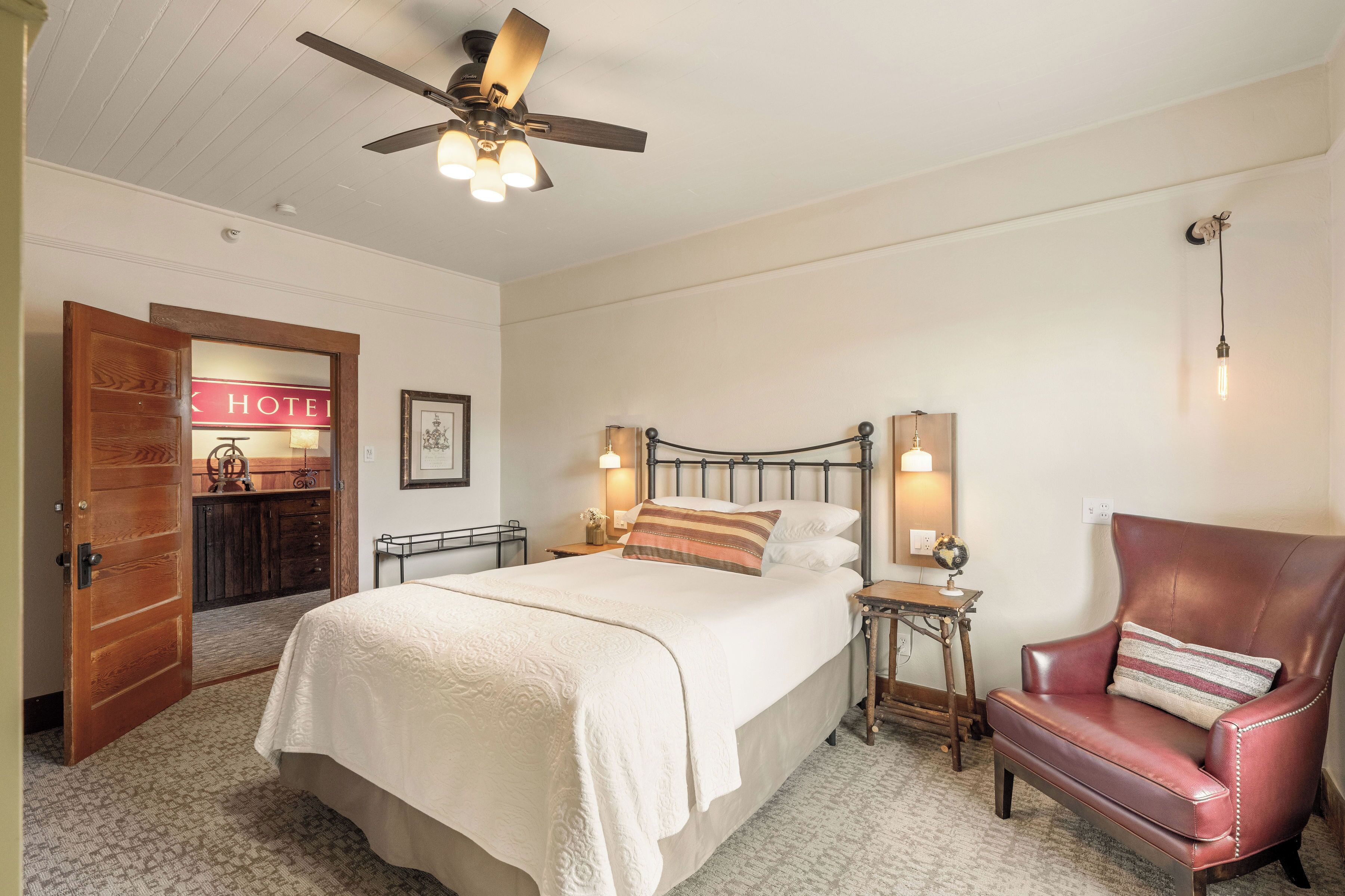A hotel room with a bed, a chair, a nightstand, a ceiling fan, and a wall-mounted light fixture.