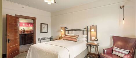 A hotel room with a bed, a chair, a nightstand, a ceiling fan, and a wall-mounted light fixture.