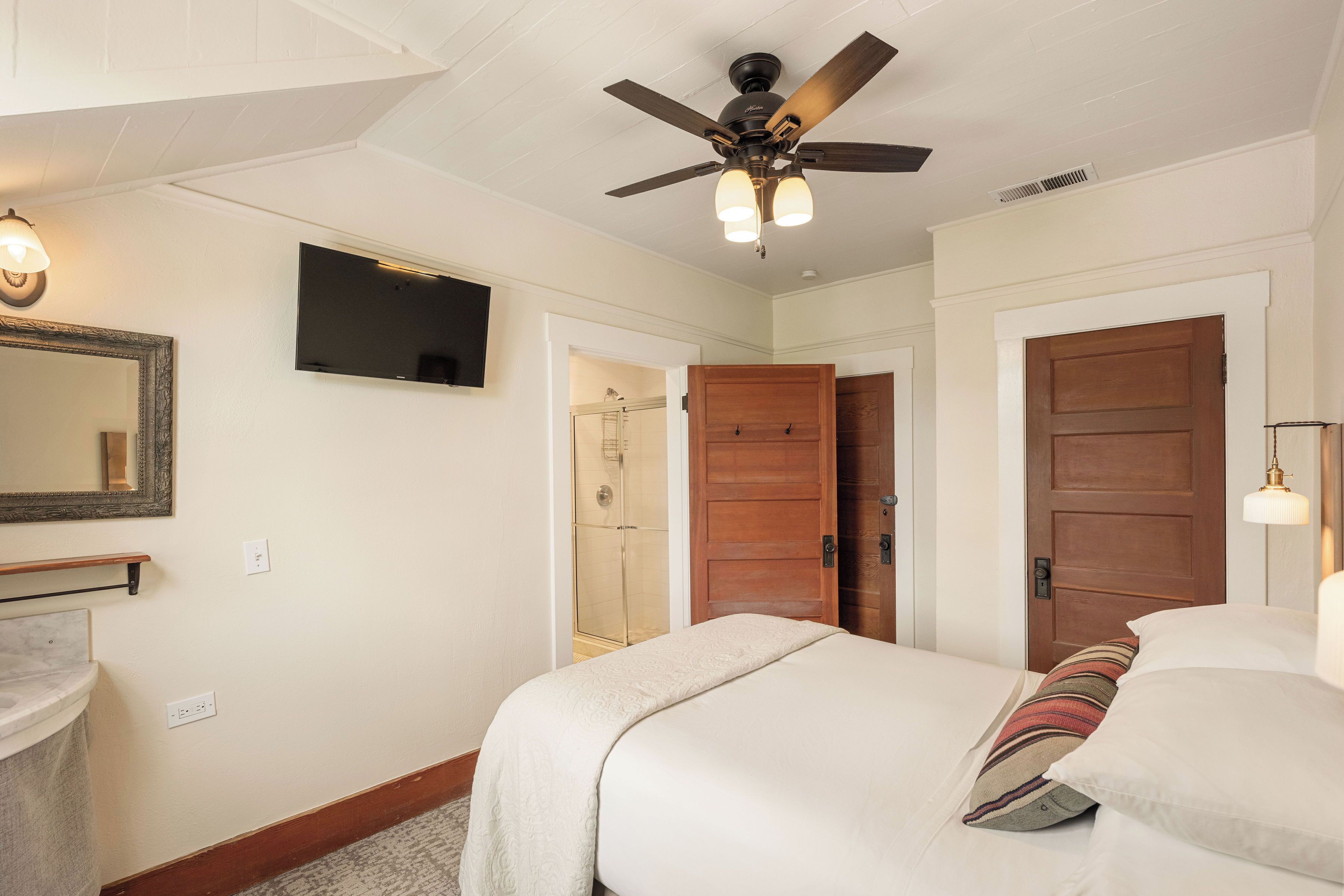 A bedroom with a bed, a ceiling fan, a TV, a wooden door, and a shower area.