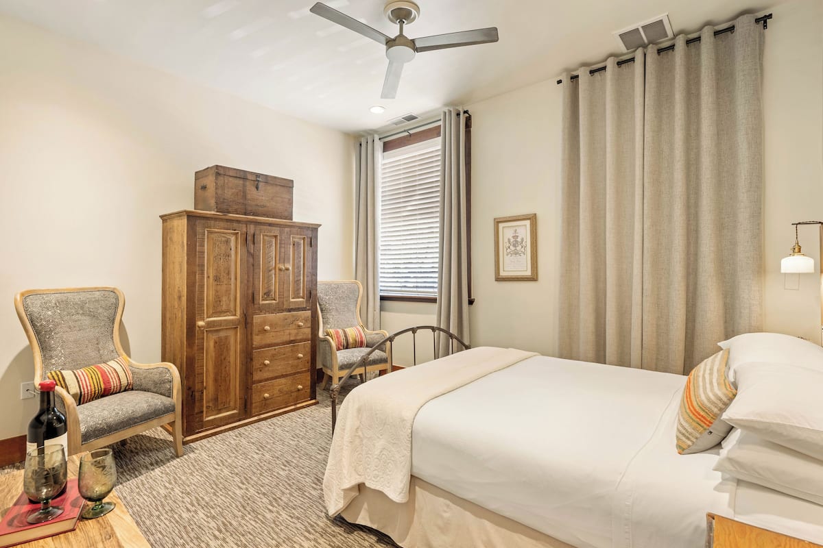 Une chambre à coucher comprenant un lit, deux chaises, une armoire en bois, un ventilateur de plafond et une fenêtre avec des stores.
