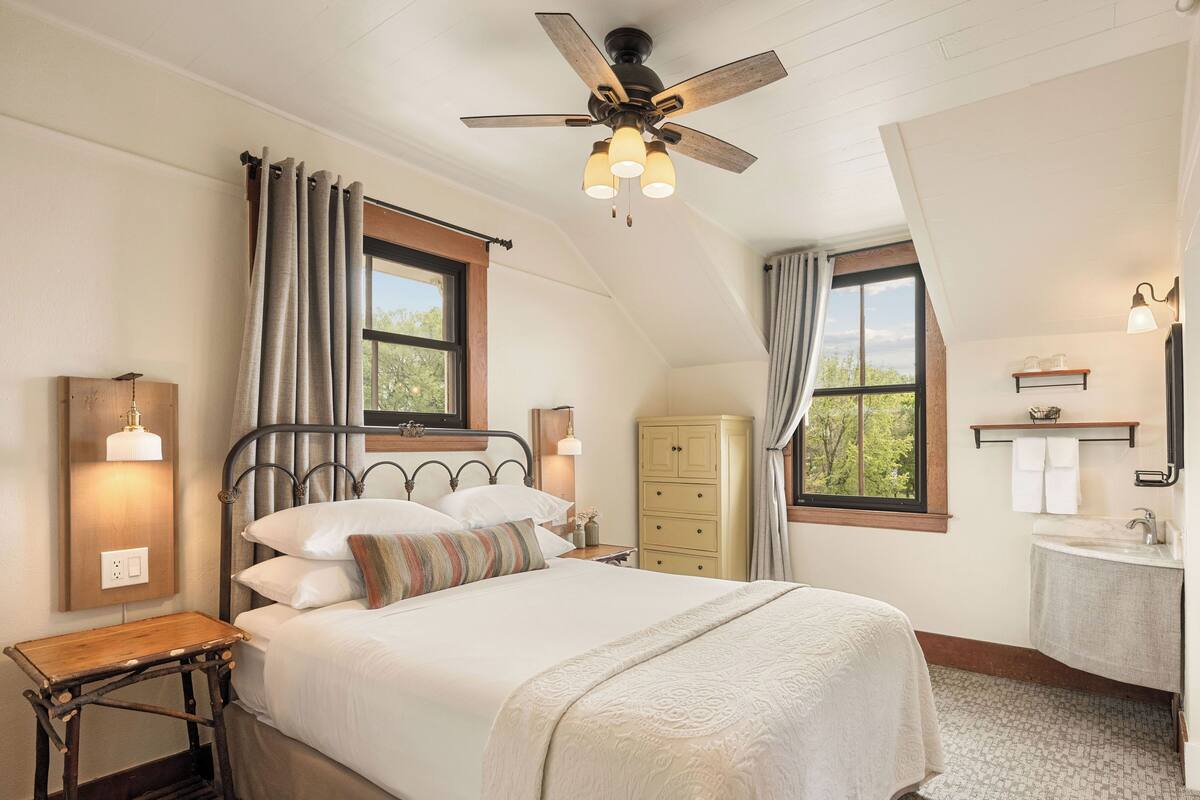 Une chambre à coucher comprenant un lit, un ventilateur de plafond, une table de chevet, une commode, une fenêtre et une salle de bain avec un lavabo et un miroir.