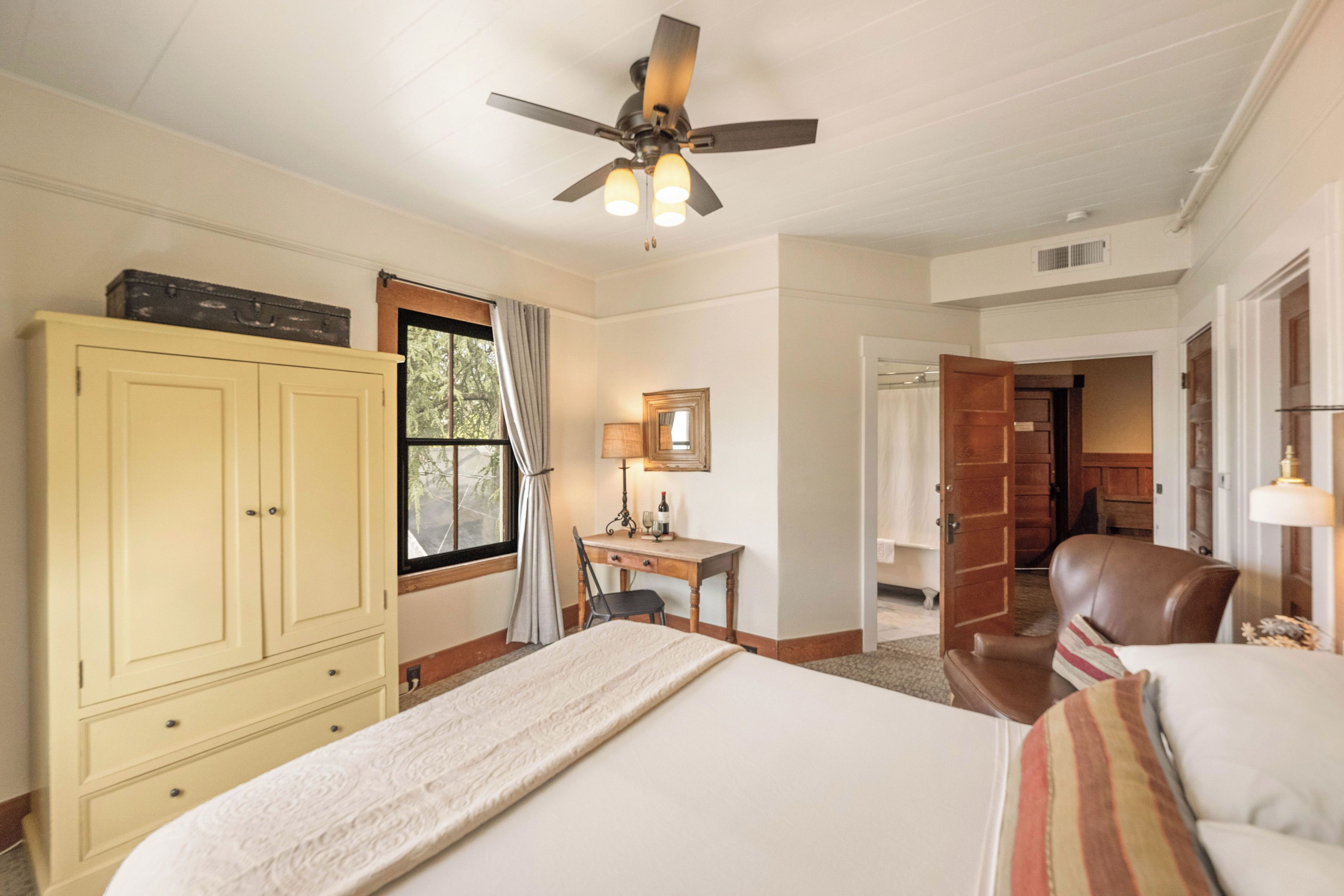 A hotel room with a bed, a desk, a chair, a wardrobe, and a ceiling fan.