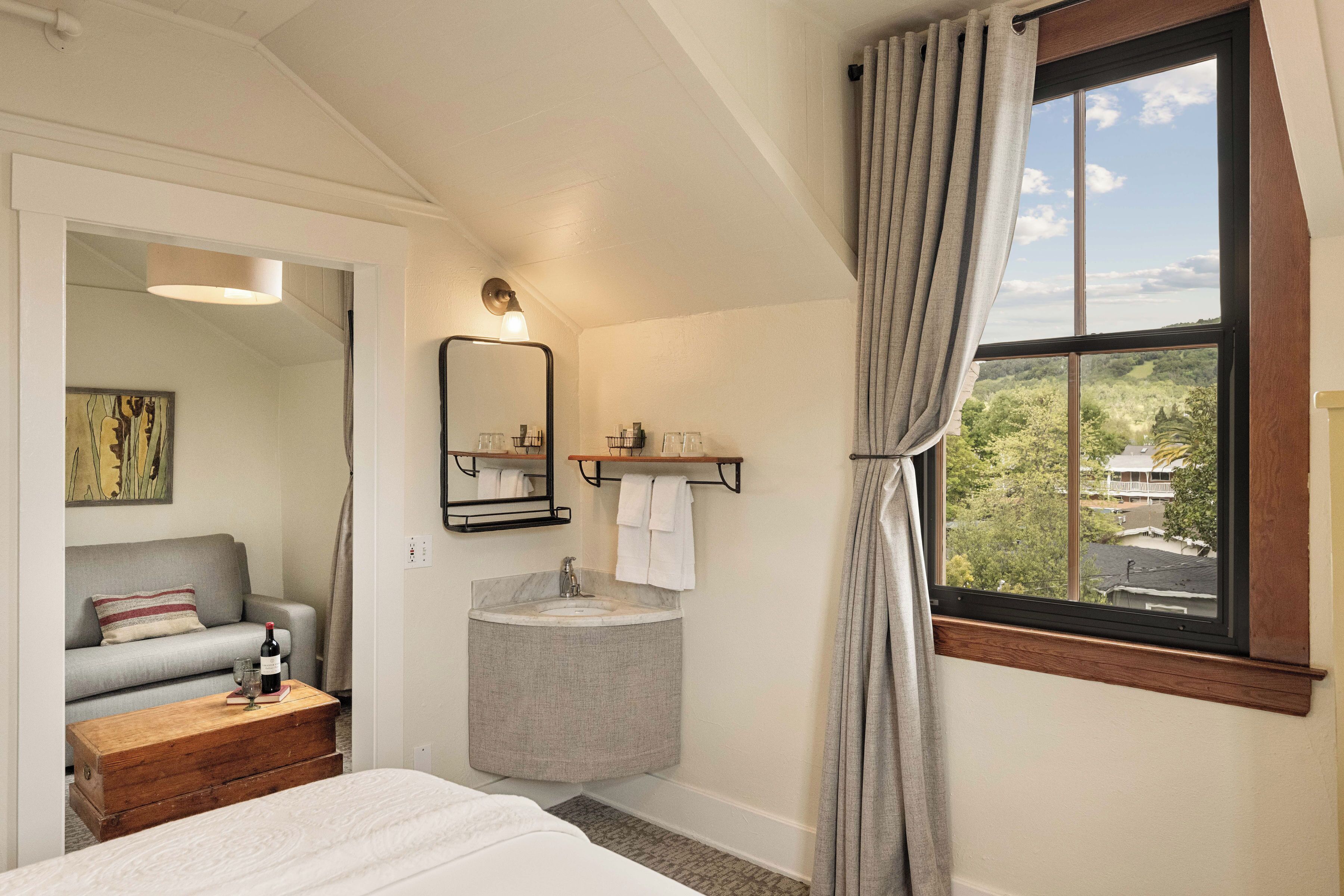 Ein Schlafzimmer mit einem Bett, einem Nachttisch aus Holz, einem runden Waschtisch, einem Spiegel und einem Fenster mit Vorhängen.
