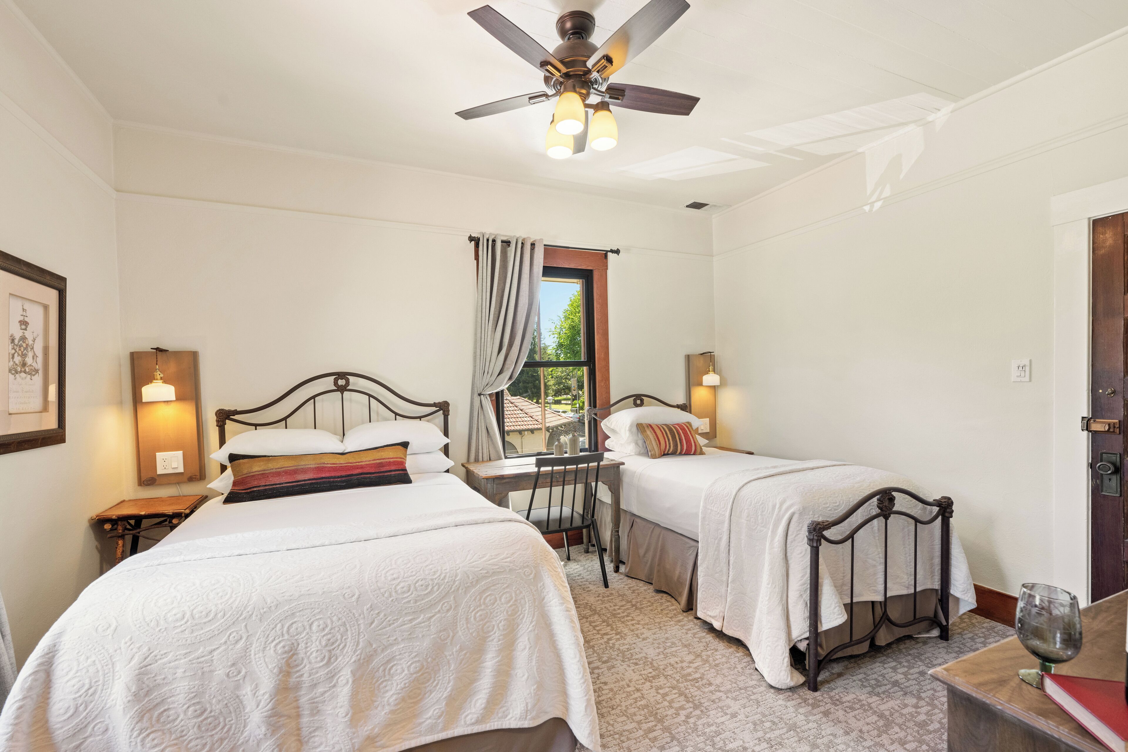 A bedroom with two single beds, a desk, a chair, a ceiling fan, and a window with curtains.