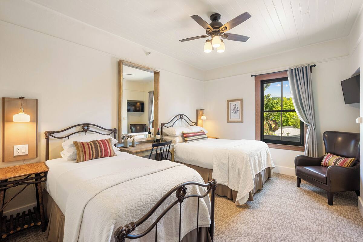 Une chambre d’hôtel avec deux lits, un ventilateur de plafond, une grande fenêtre avec vue, un bureau avec une chaise et un téléviseur fixé au mur.