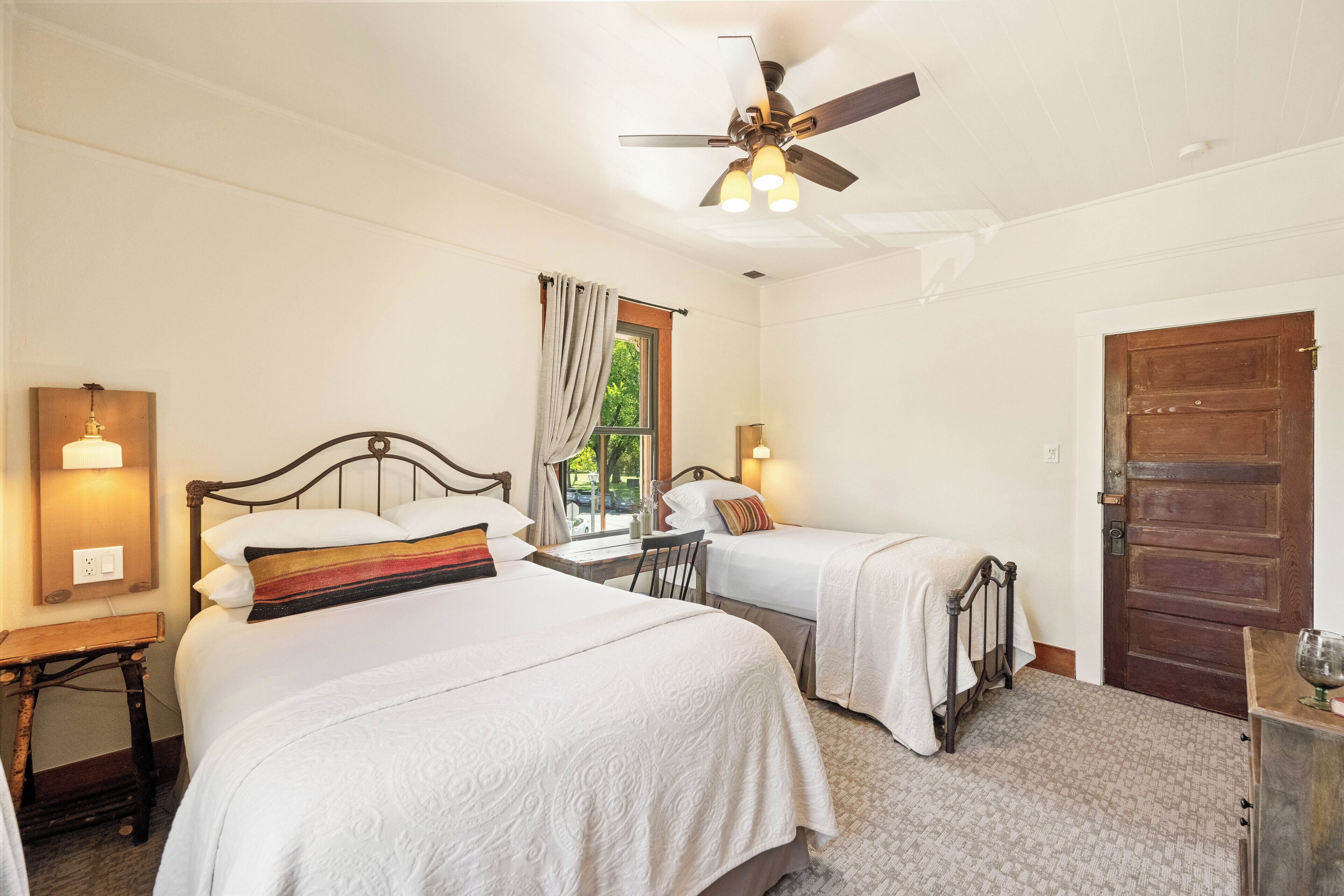 A hotel room with two beds, a ceiling fan, a window with curtains, a wooden door, and a small table with a lamp.