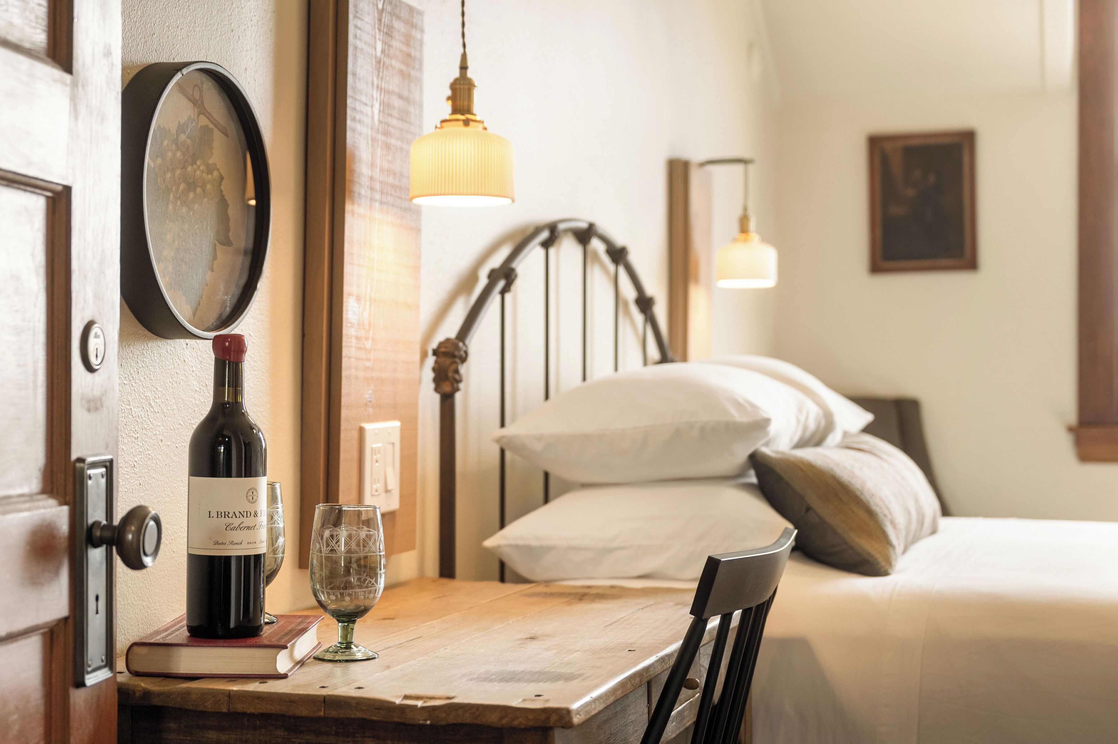 A room with a wooden table, a wine bottle, and a glass on it. There is a bed with pillows and a headboard in the background.