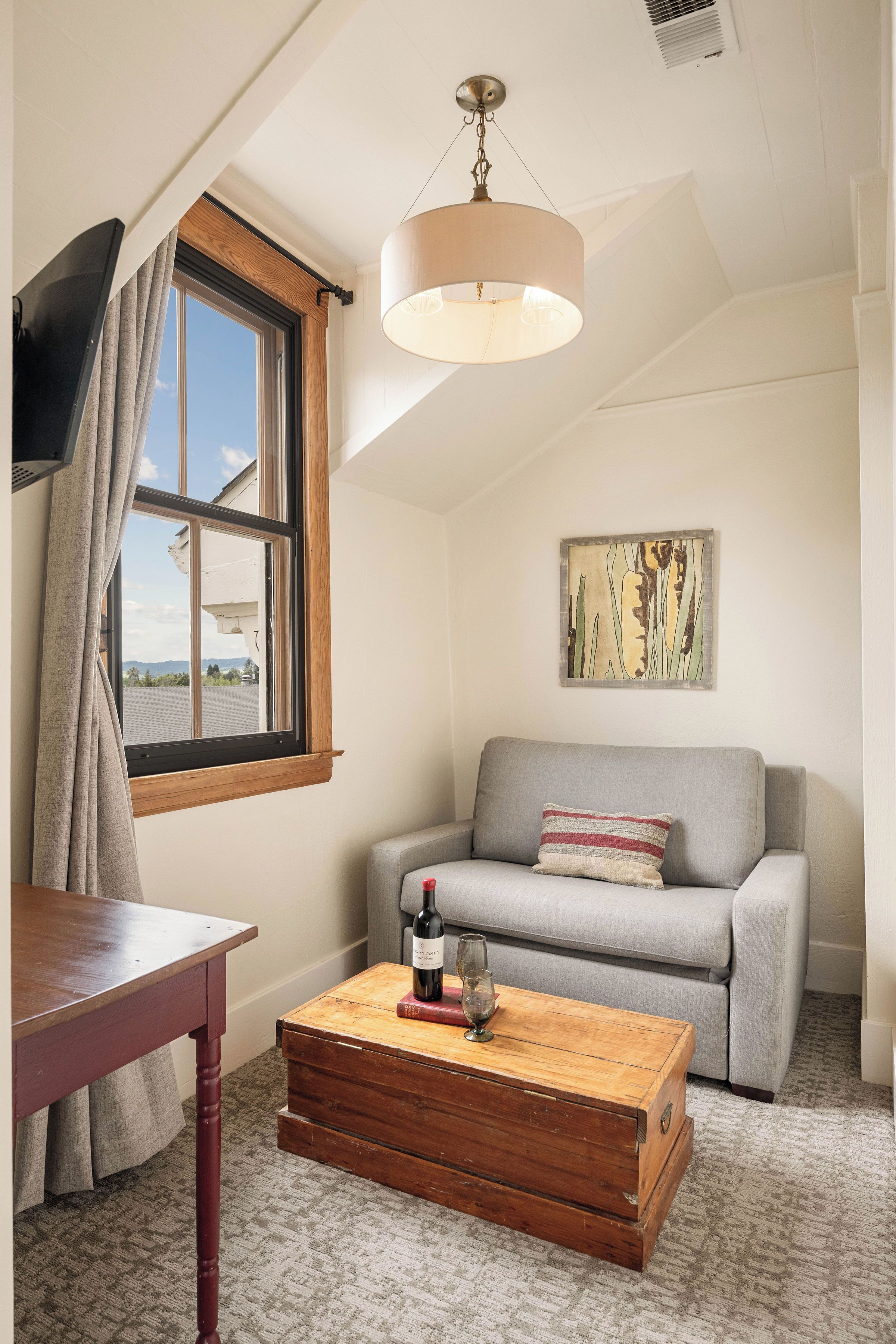 Una habitación acogedora con una mesa de madera, un sofá gris, un aparador de madera, una lámpara colgante y una ventana con cortinas.