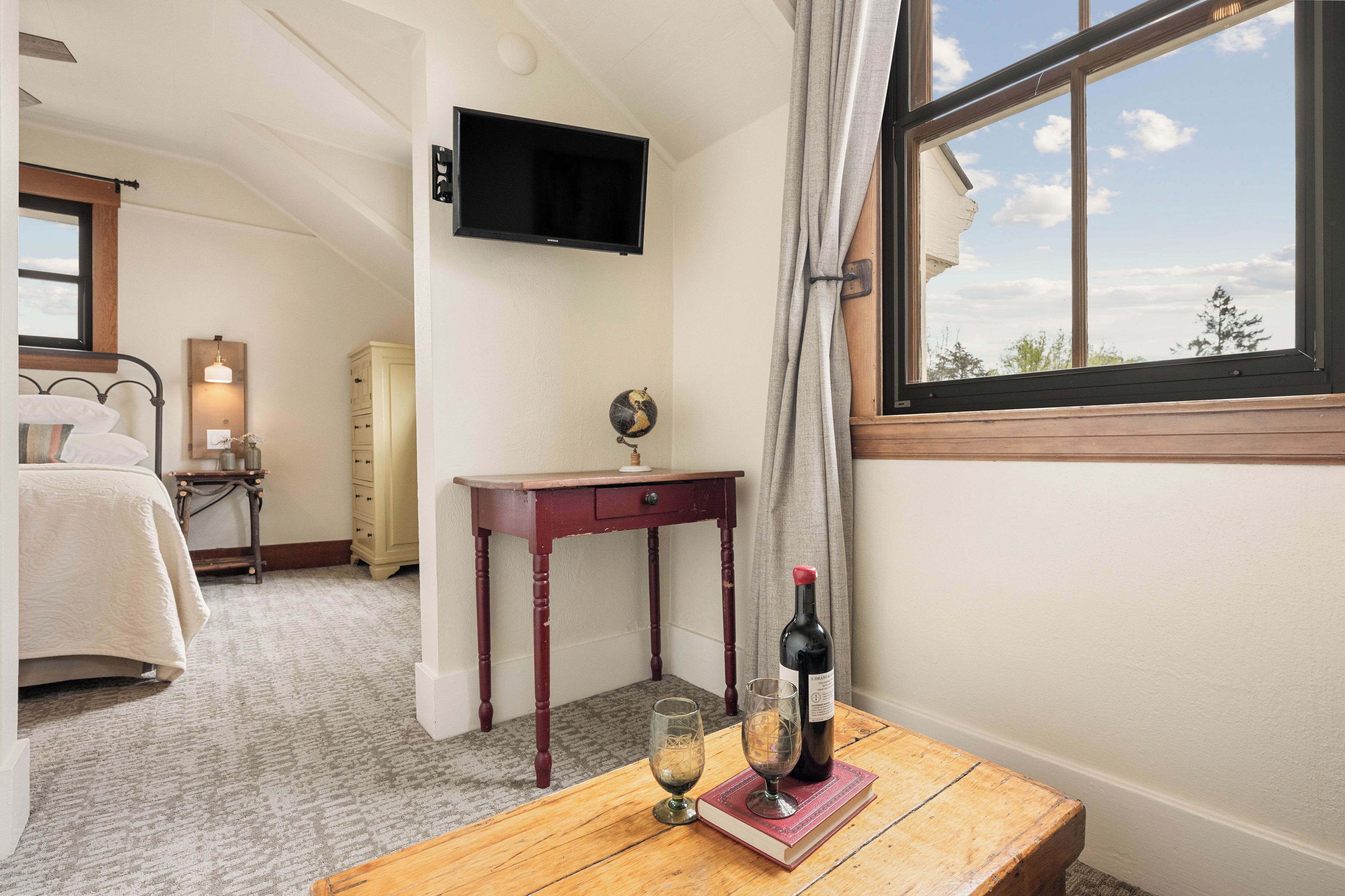 Un dormitorio con cama, mesita de noche con botella de vino y copa, televisor montado en la pared y ventana con cortinas.