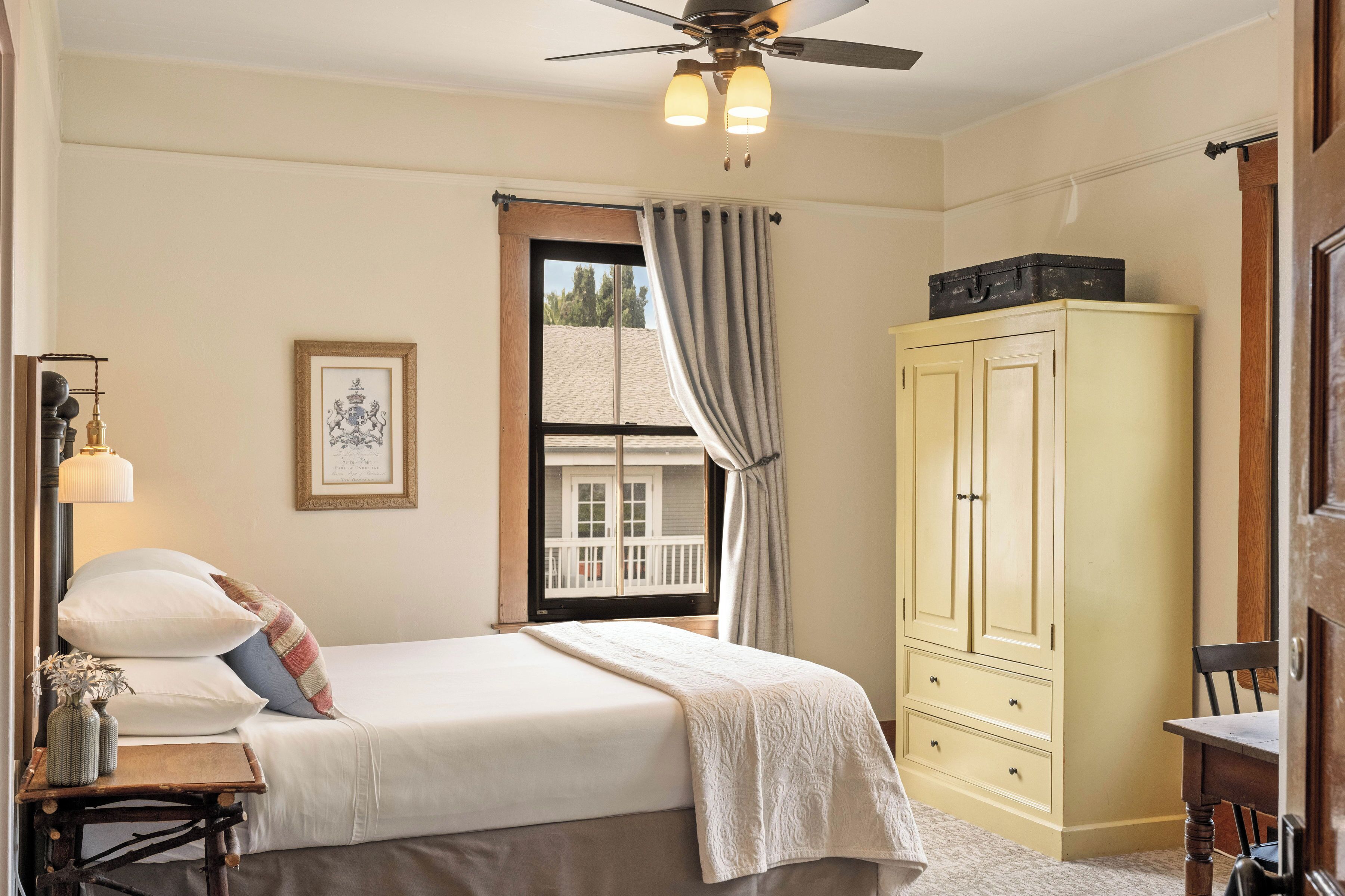 A bedroom with a bed, a wardrobe, a desk, a lamp, and a window with curtains.