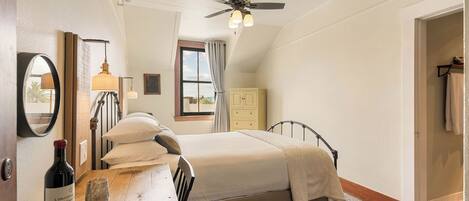 A bedroom with a bed, a bedside table with a wine bottle and glass, a ceiling fan, a window with curtains, and a wardrobe.