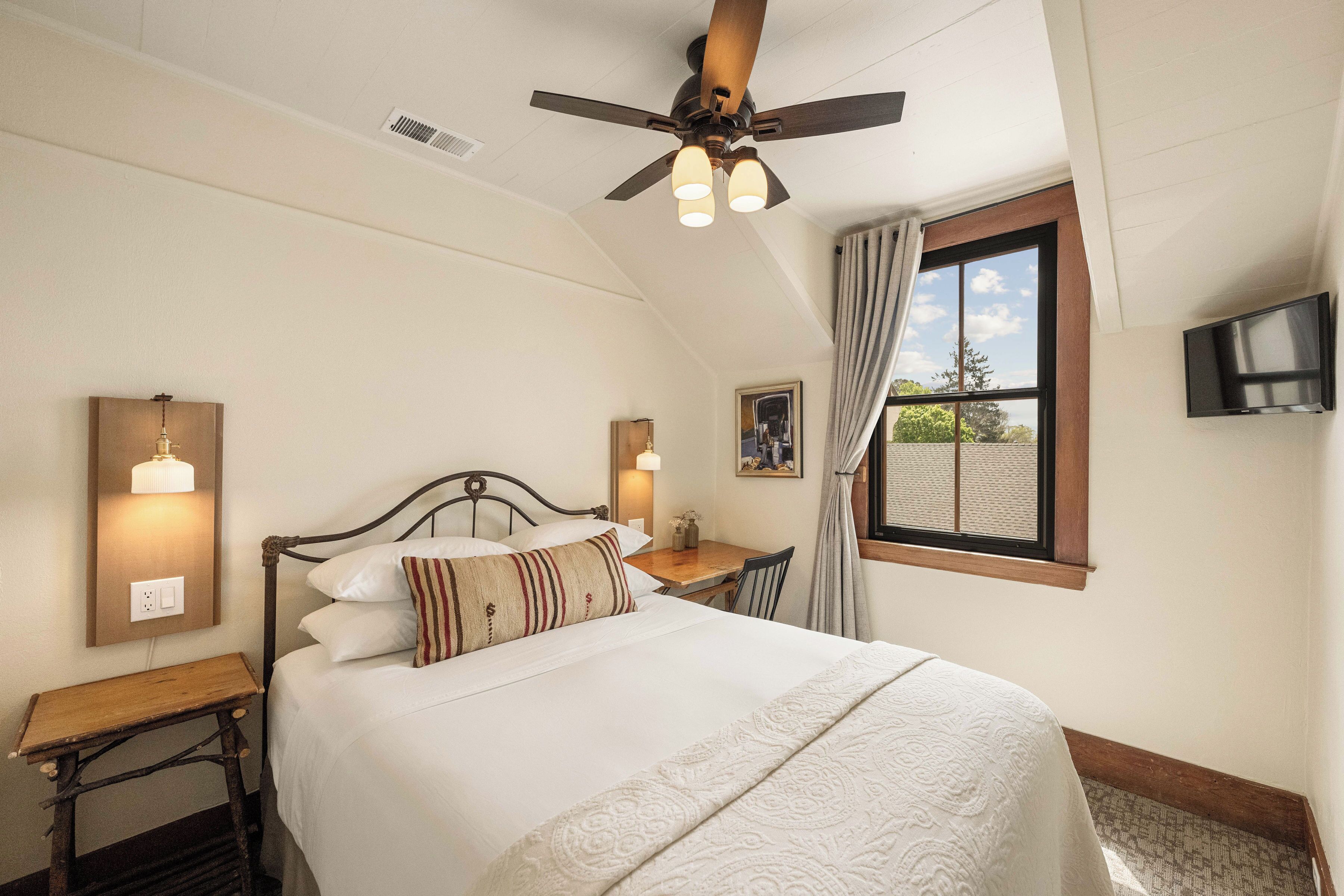 A bedroom with a bed, bedside table, ceiling fan, window, and TV.