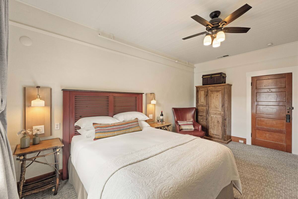 Une chambre d’hôtel comprenant un lit, une armoire en bois, une chaise, un bureau avec une lampe et un ventilateur de plafond.