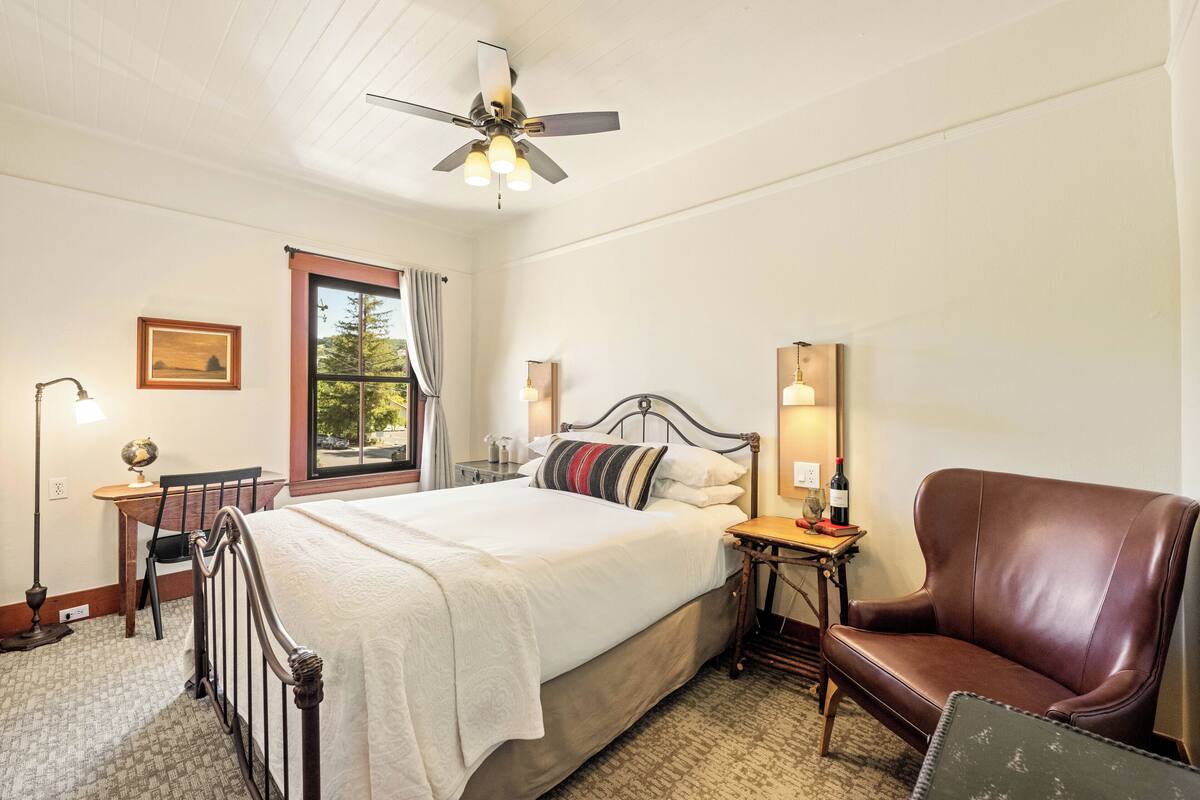 Une chambre à coucher comprenant un lit, un bureau, une chaise, un fauteuil en cuir, un ventilateur de plafond et une fenêtre avec des rideaux.