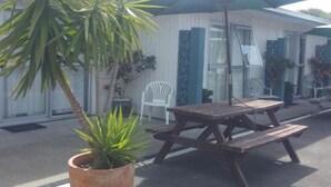 Un patio con mesa y bancos de madera, rodeado de plantas en macetas y una silla blanca.