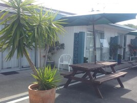 Un patio con mesa y bancos de madera, rodeado de plantas en macetas y una silla blanca.