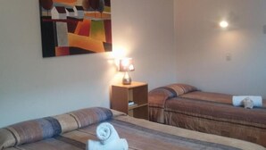 Habitación de hotel con dos camas, un cuadro en la pared, una mesita de noche con lámpara y un estante pequeño.