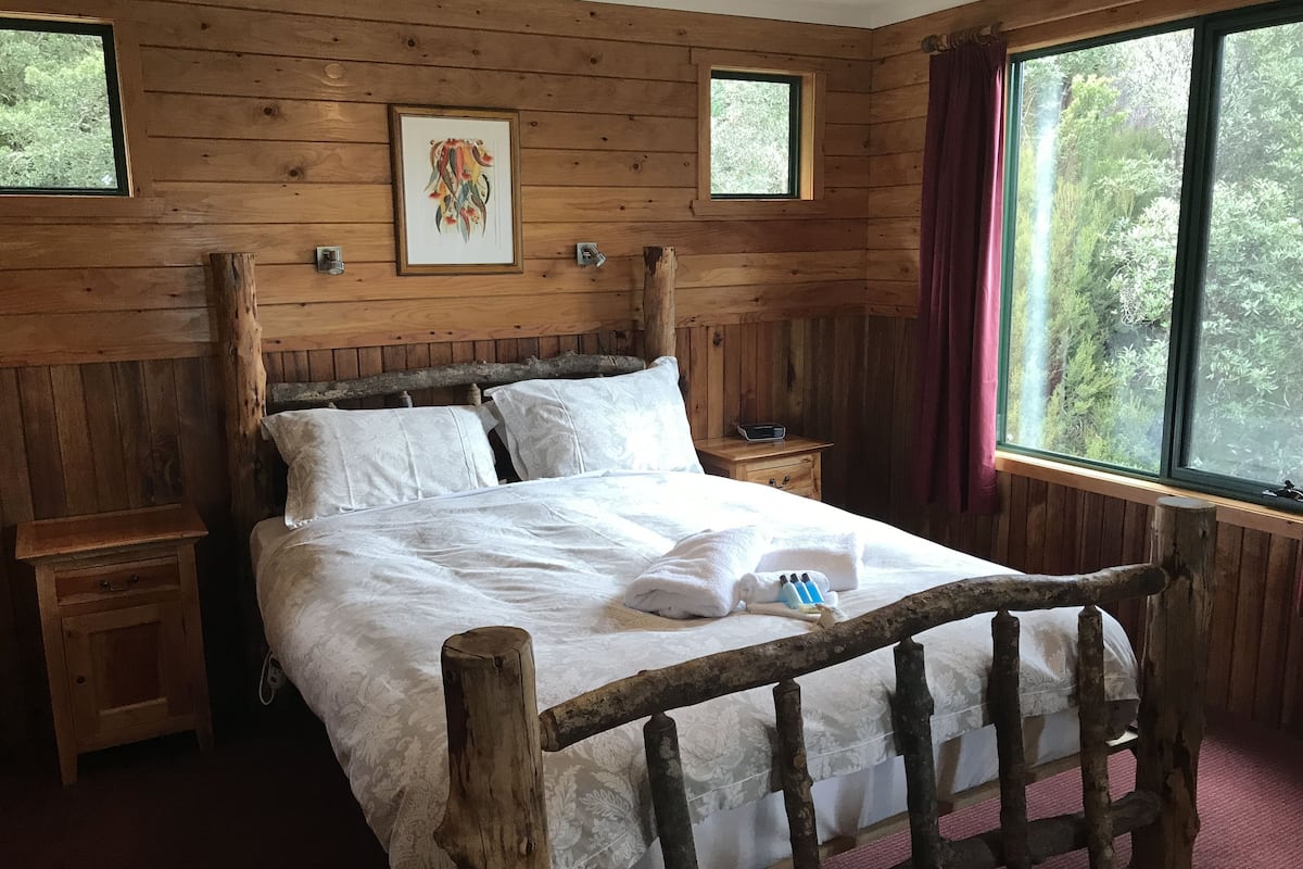Una cama con estructura de madera y ropa de cama blanca, una mesita de noche con una radio y una ventana que da a vistas de árboles.