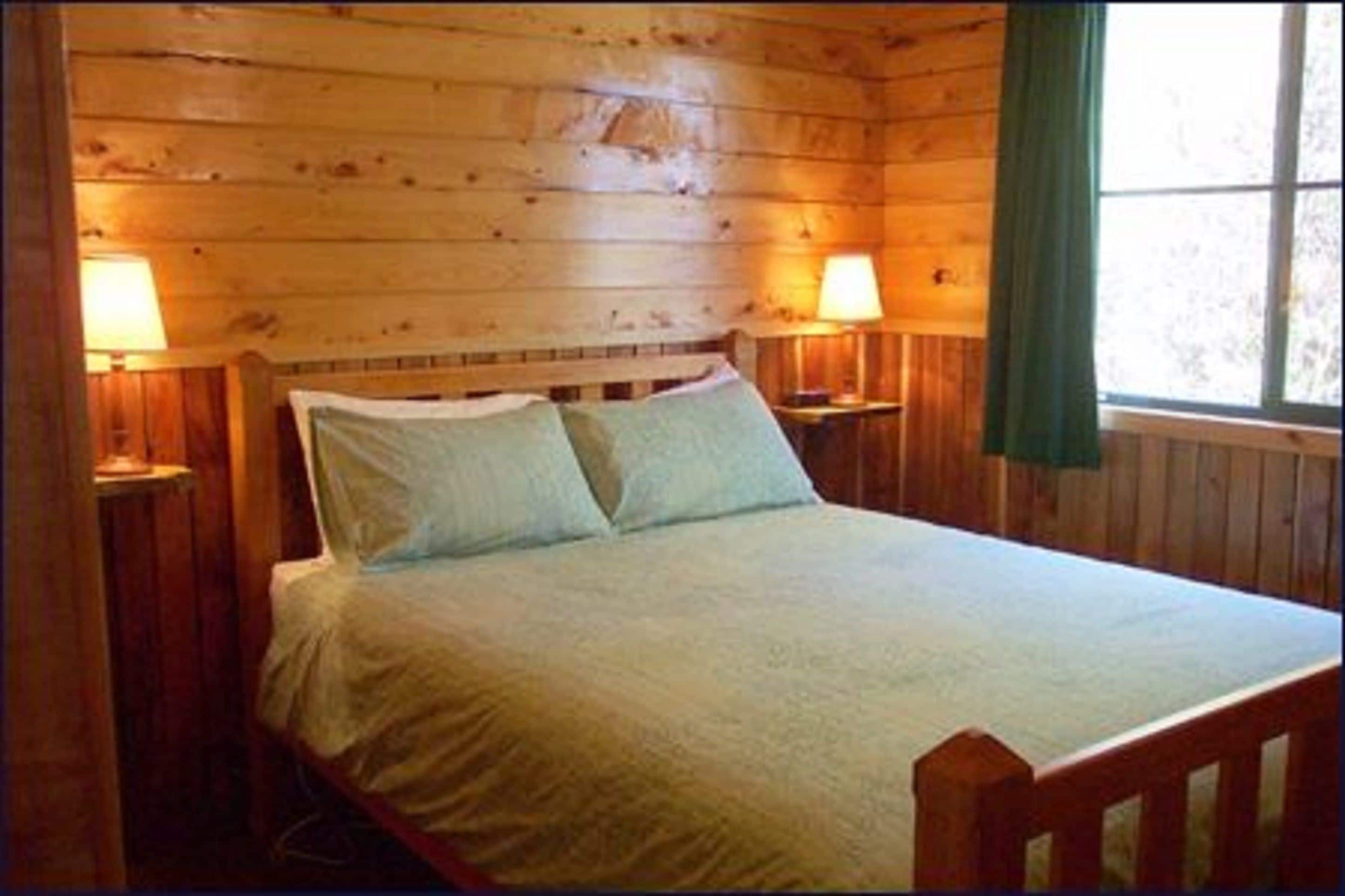 A wooden cabin-style room with a bed, two bedside lamps, and a window with curtains.