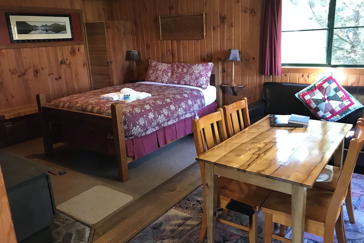 Habitación con paneles de madera que incluye una cama, una mesa y una silla. Se observa una ventana con cortinas, una lámpara y un cuadro enmarcado en la pared.
