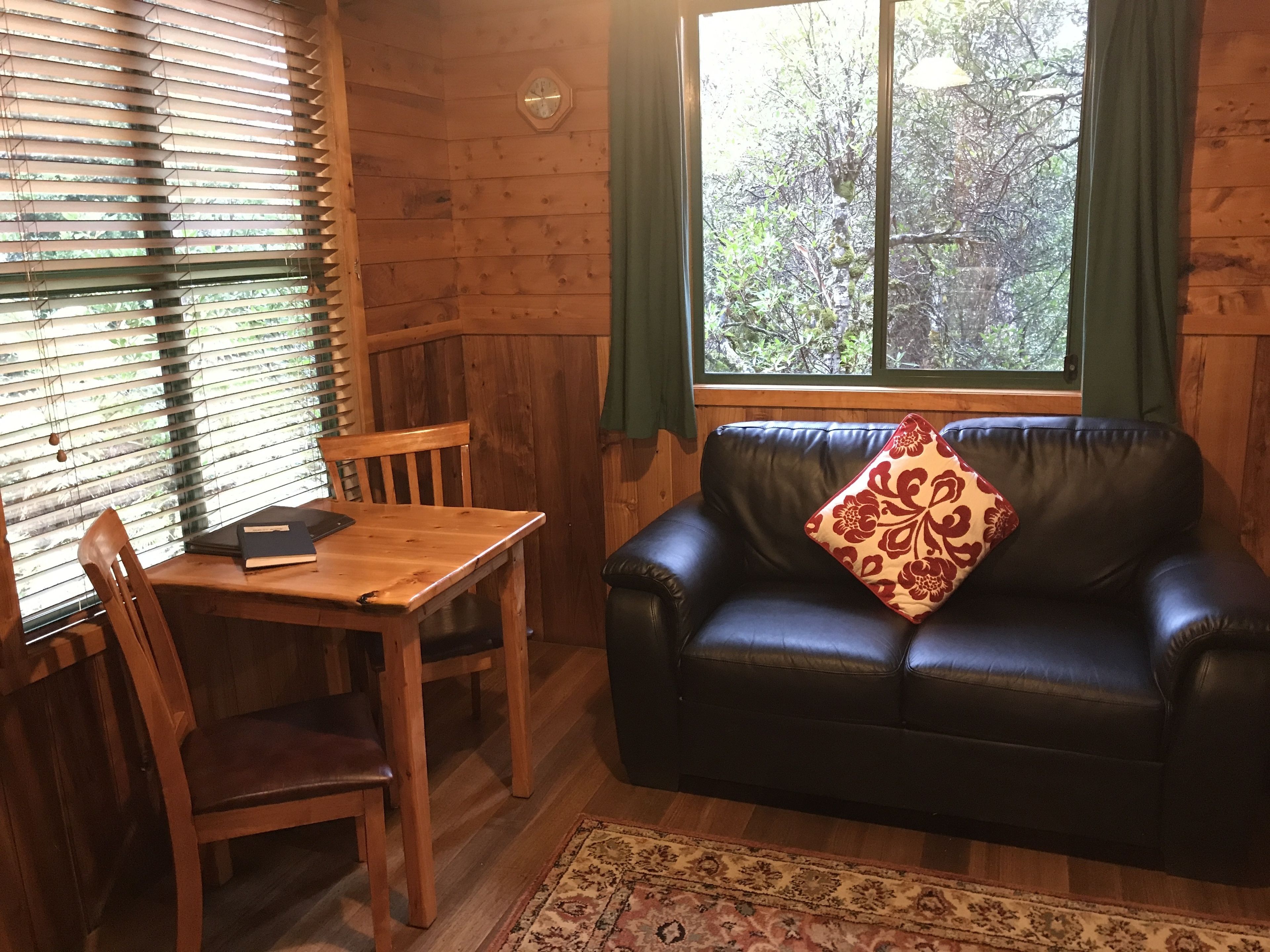 Una habitación acogedora con una mesa de madera, un sofá de cuero y un cojín floral, además de un amplio ventanal con cortinas.